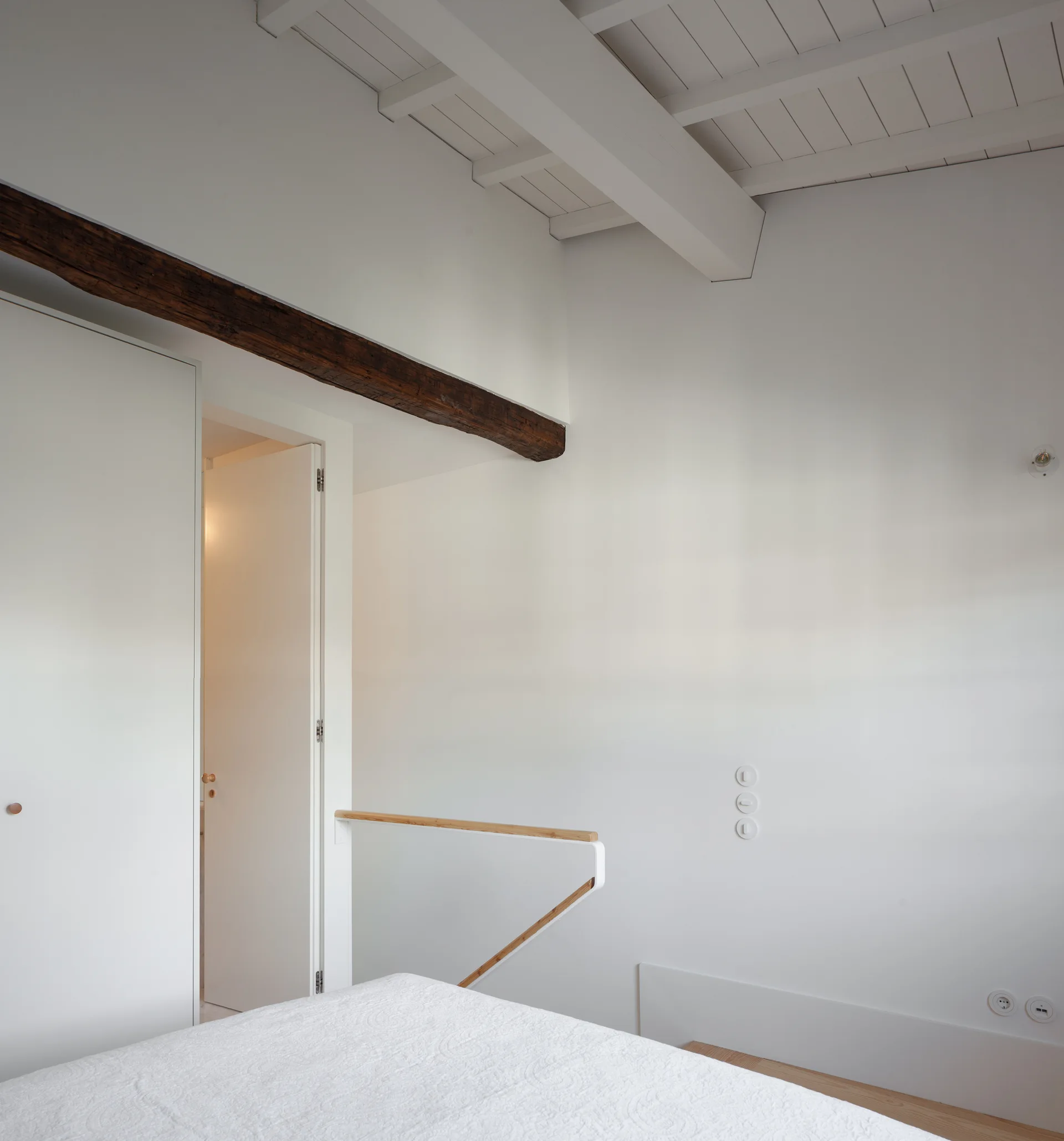 A minimalist white loft bedroom features a prominent dark wood beam on the wall and a light wood stair railing.