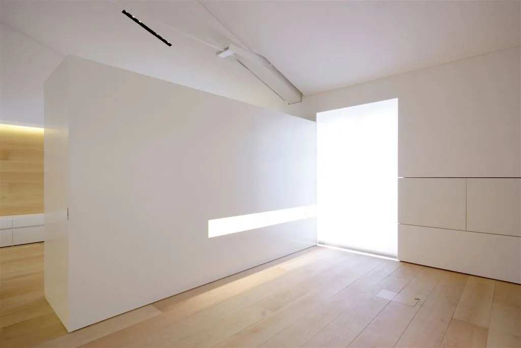 A minimalist room features a large white partition wall with a glowing horizontal light strip, pale wood floors, and seamless cabinetry.
