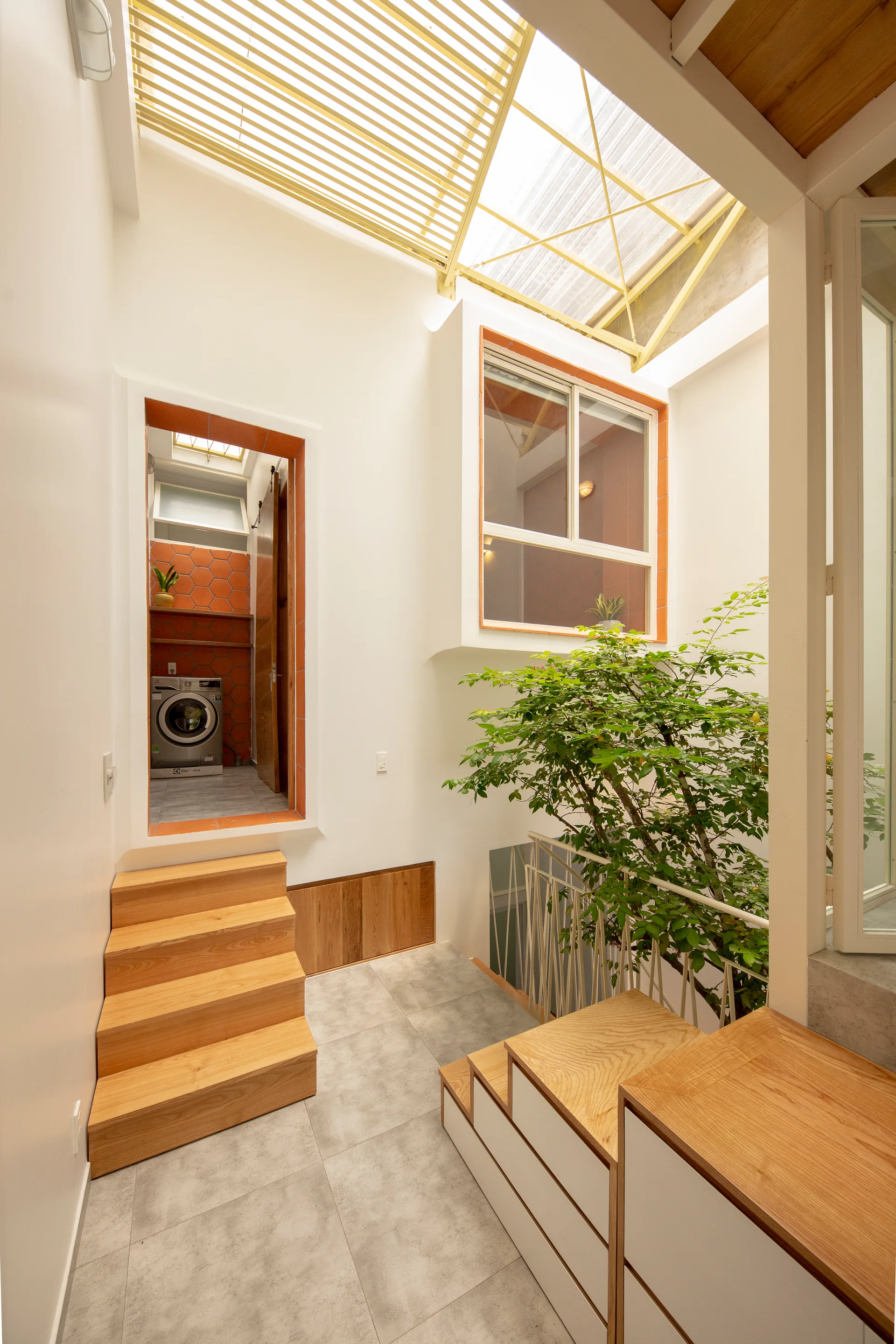 A bright residential stairwell features wooden steps, a central green tree on a landing, and a skylight with yellow slats above.