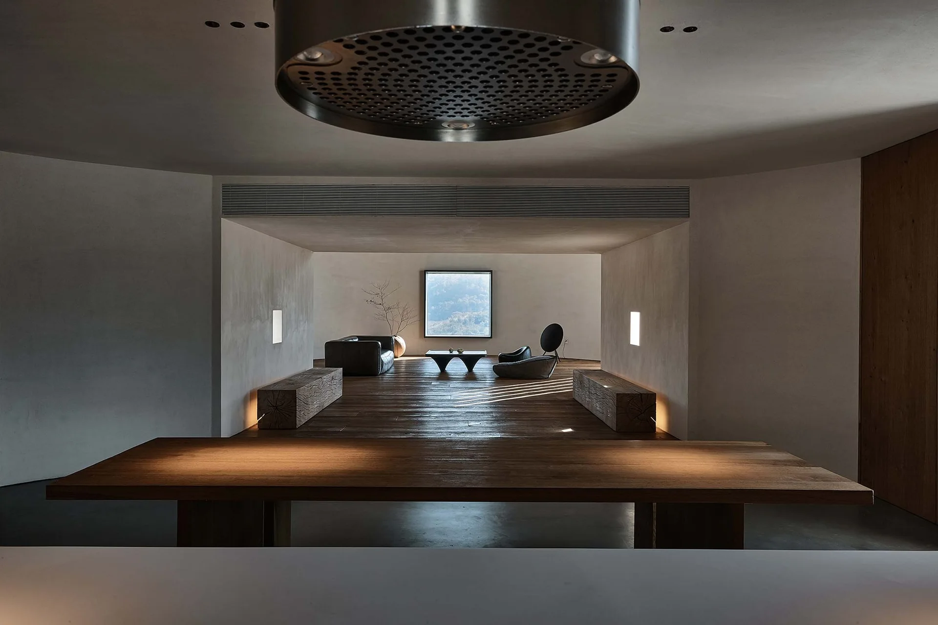 A minimalist living room features raw wood block benches, low-profile dark furniture, plaster walls, and a square window framing a nature view.