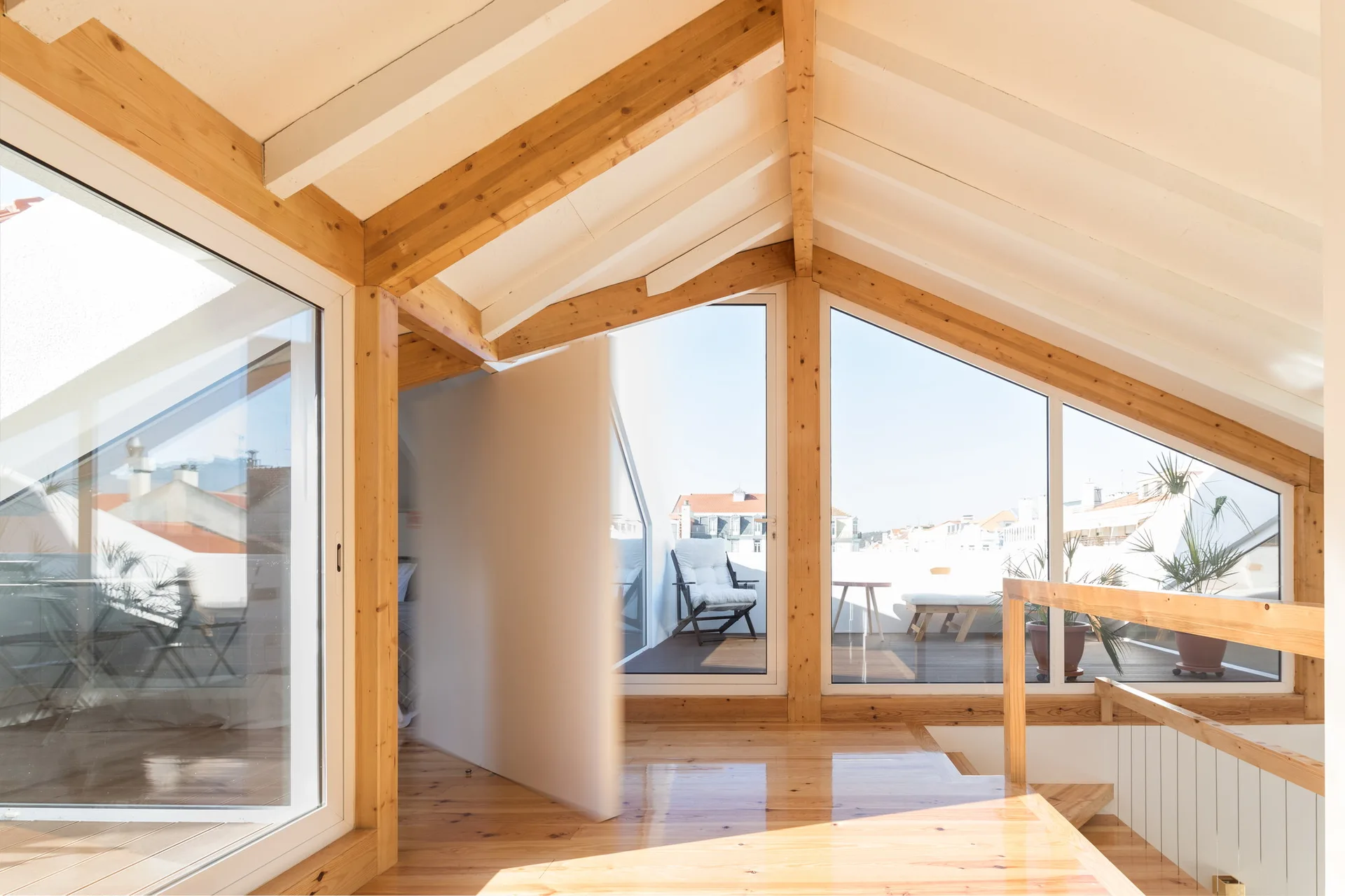 An attic landing features polished wood floors, exposed timber ceiling beams, and large angled windows leading to a sunny outdoor terrace.
