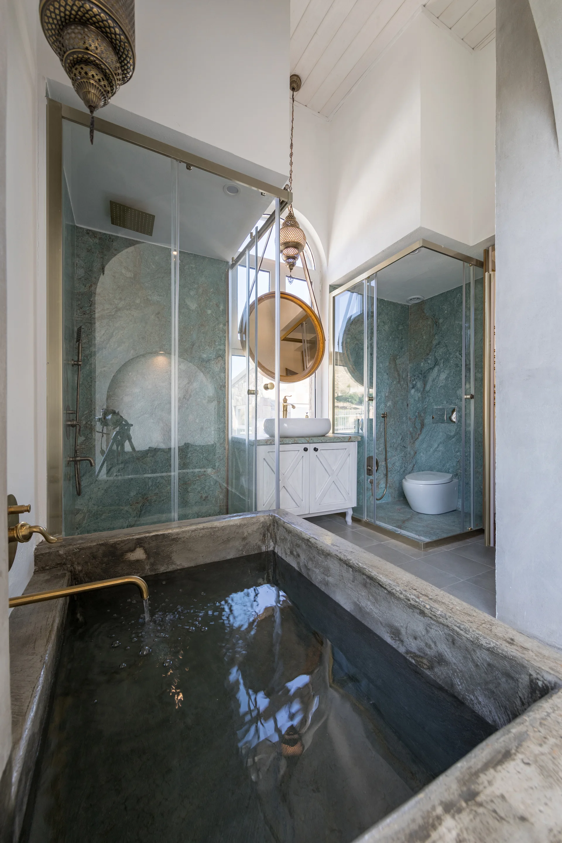 A bathroom with a raw concrete soaking tub in the foreground, green marble walls in glass-enclosed stalls, and a white vanity cabinet.