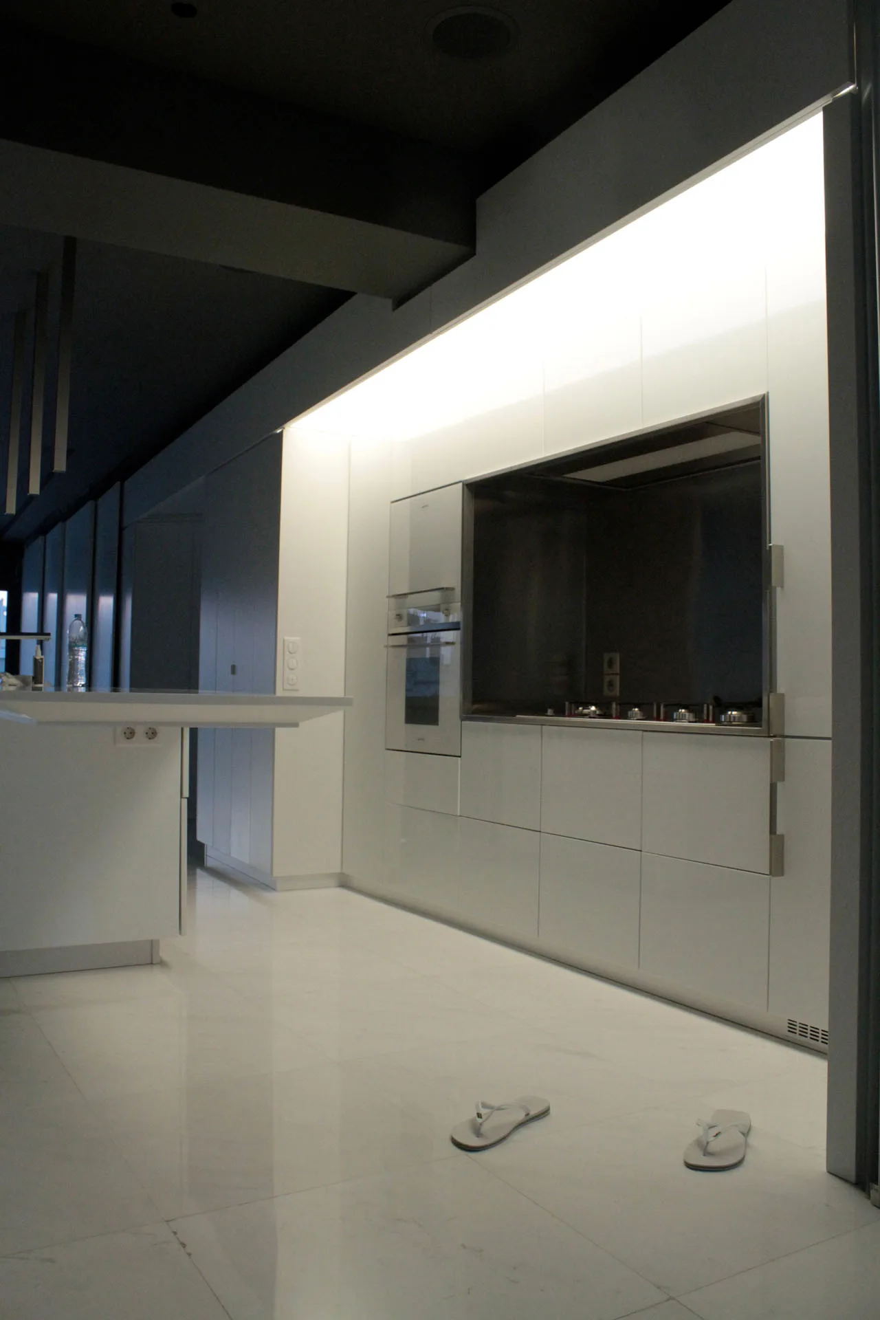 A minimalist white kitchen features flat-panel glossy cabinets, integrated appliances, large-format tile flooring, and a dark ceiling with soft cove lighting.