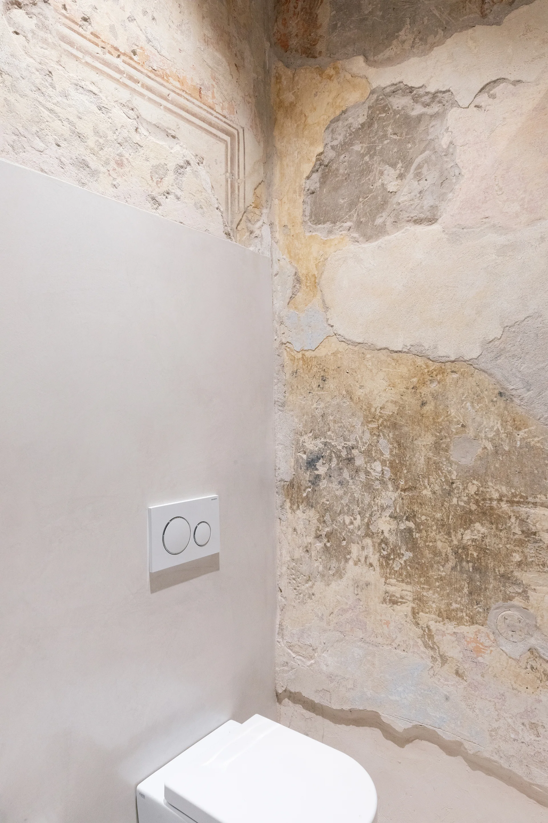 A modern white toilet and flush plate mounted on a smooth gray panel next to a weathered, distressed plaster wall in a bathroom.