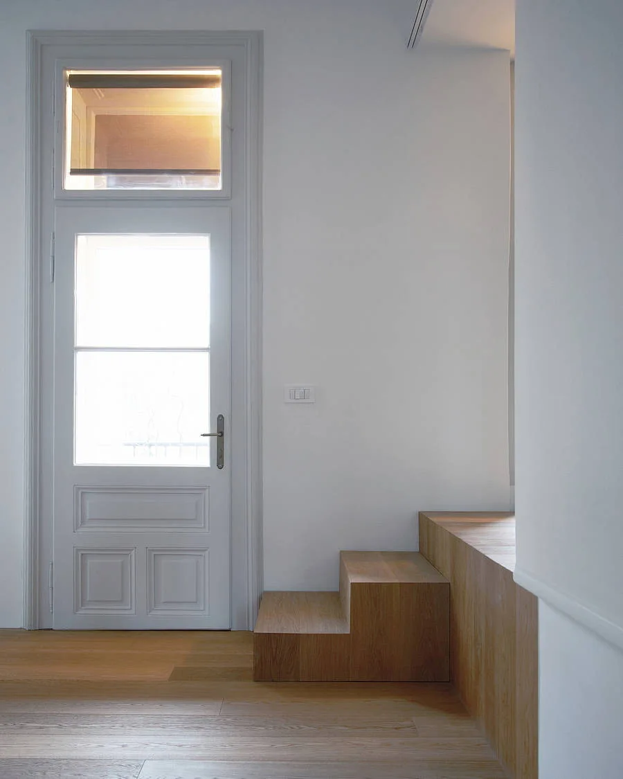 A minimalist entryway features a light wood floor, a tall light gray door with a glass pane, and a matching tiered wooden step platform.