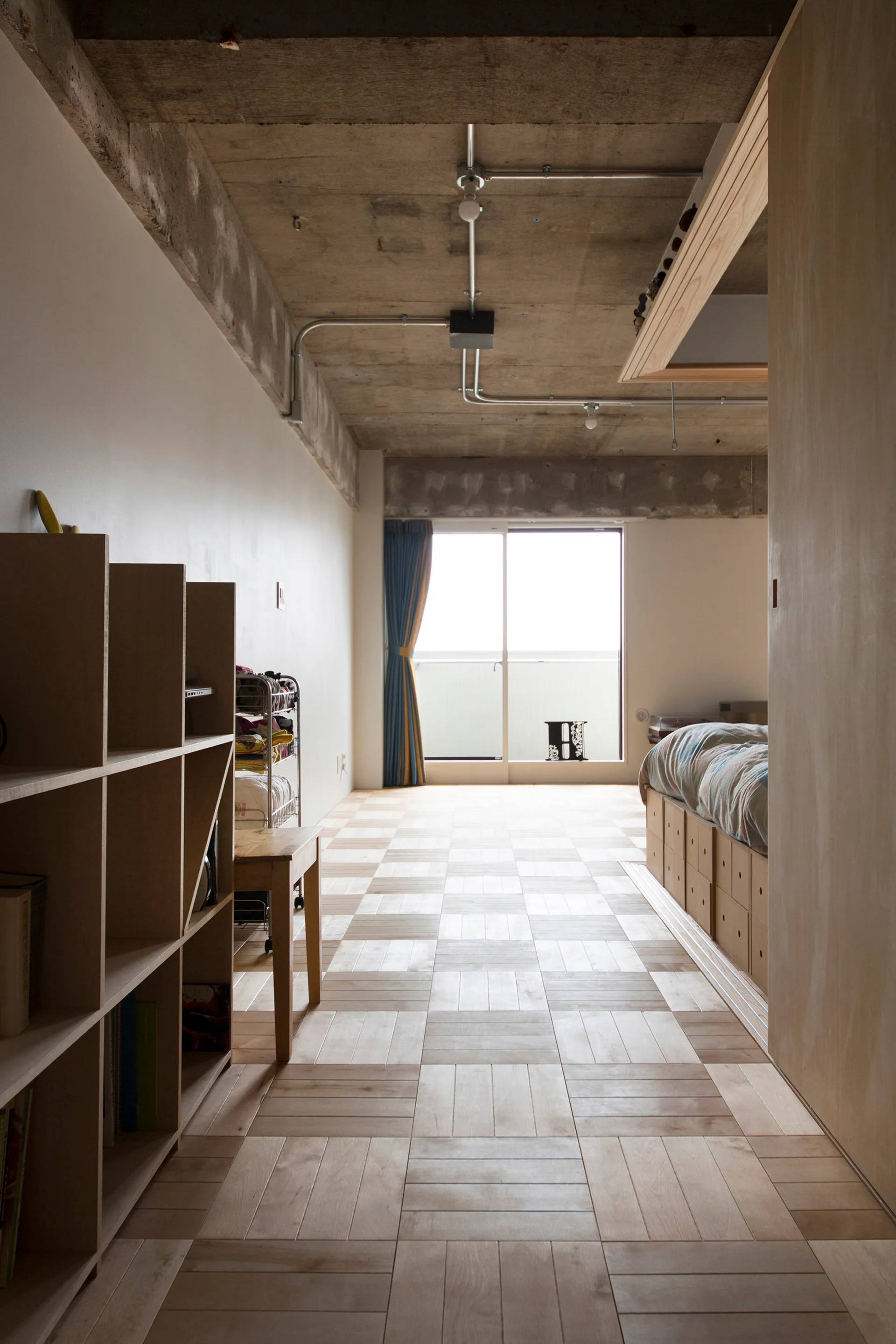 A minimalist bedroom featuring an exposed concrete ceiling, light wood parquet floors, a wooden platform storage bed, and a modular bookshelf.