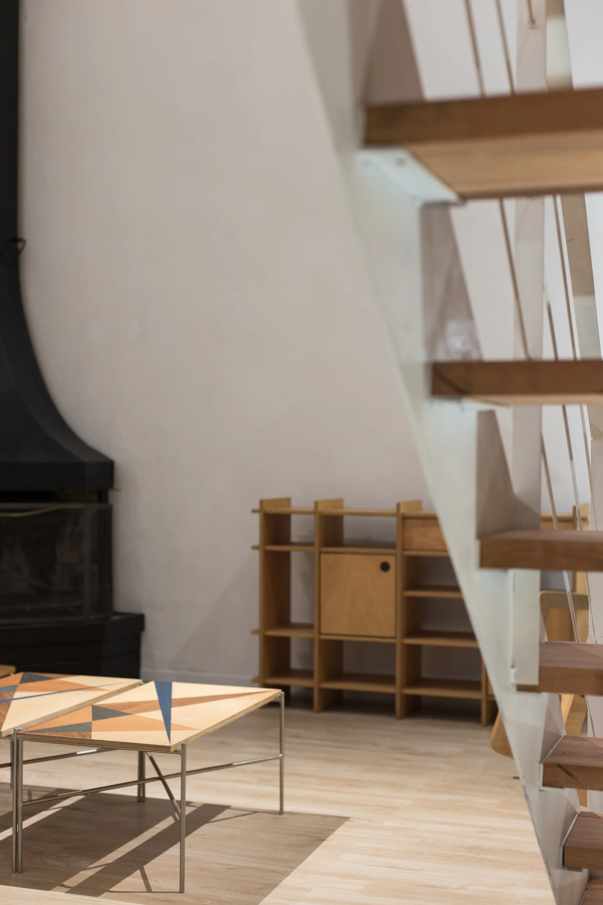 A minimalist living space featuring a wooden open-tread staircase, geometric patterned coffee tables, a light wood shelving unit, and a fireplace.