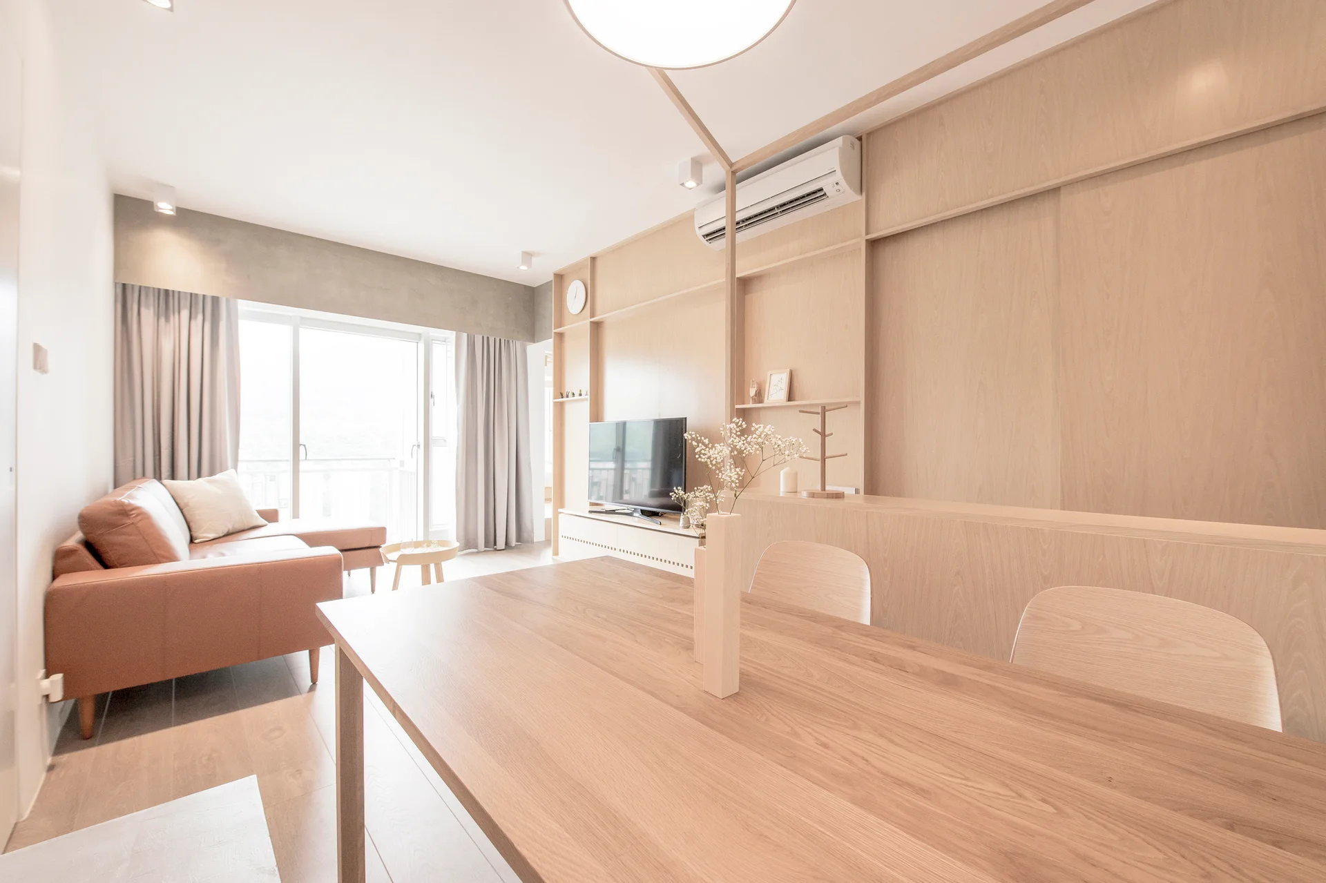An open living and dining space features a wooden dining table, a tan leather sofa, and light wood wall paneling with integrated shelving.