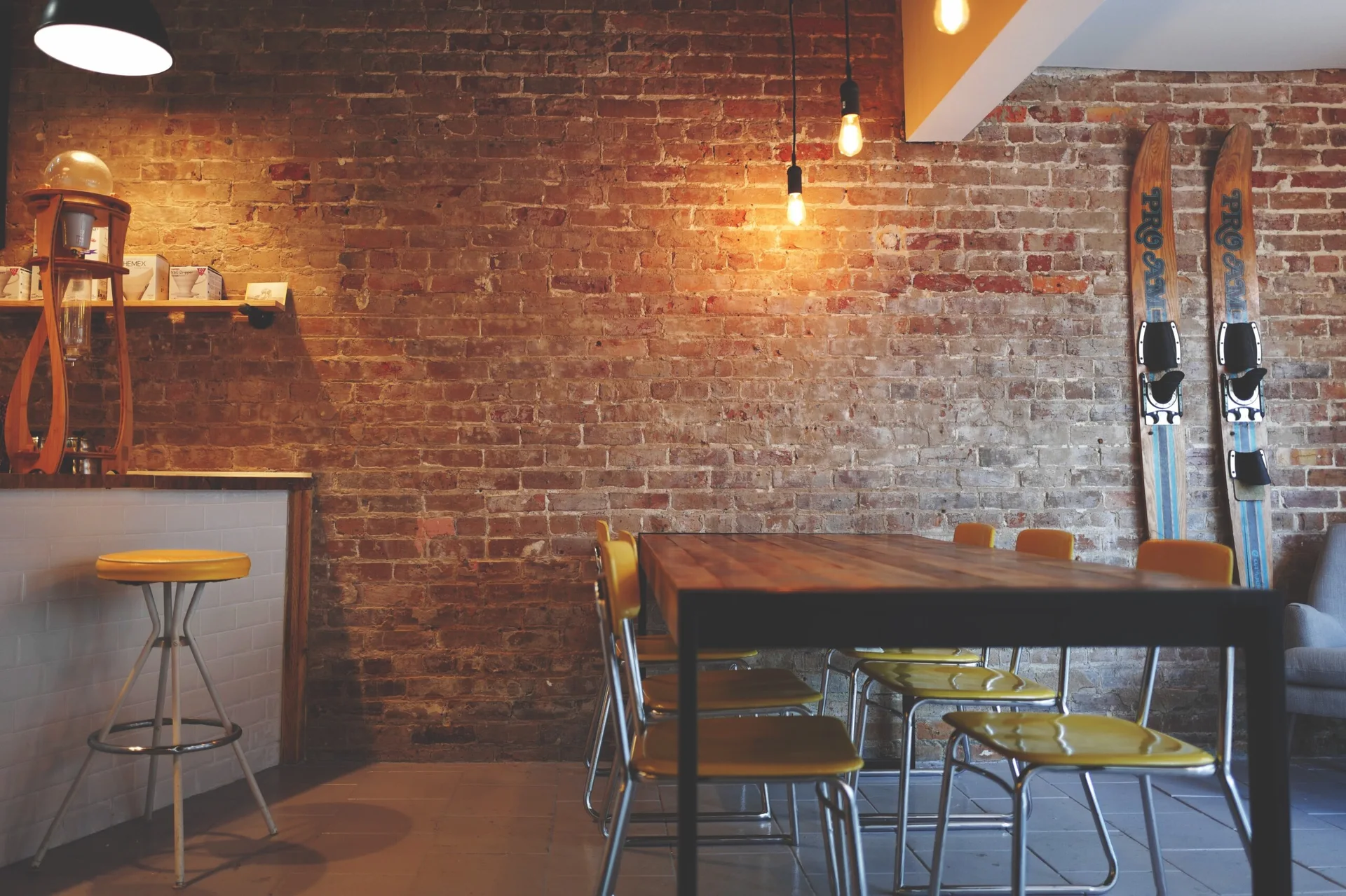 A rustic wooden dining table surrounded by yellow chairs sits in front of an exposed brick wall decorated with vintage wooden skis.