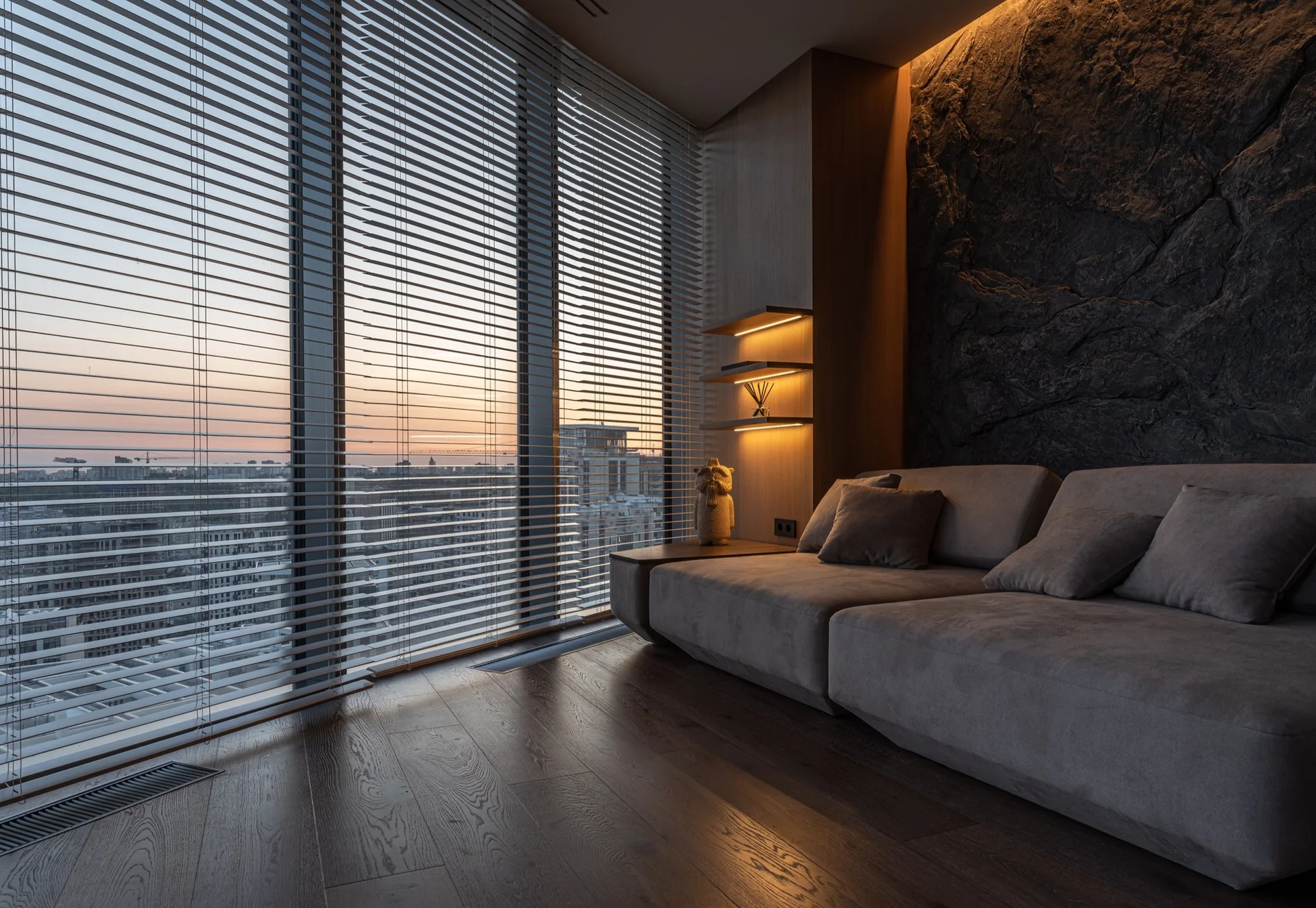 A low-profile grey sofa sits in front of a dark textured stone wall next to lighted shelves and a window with venetian blinds.