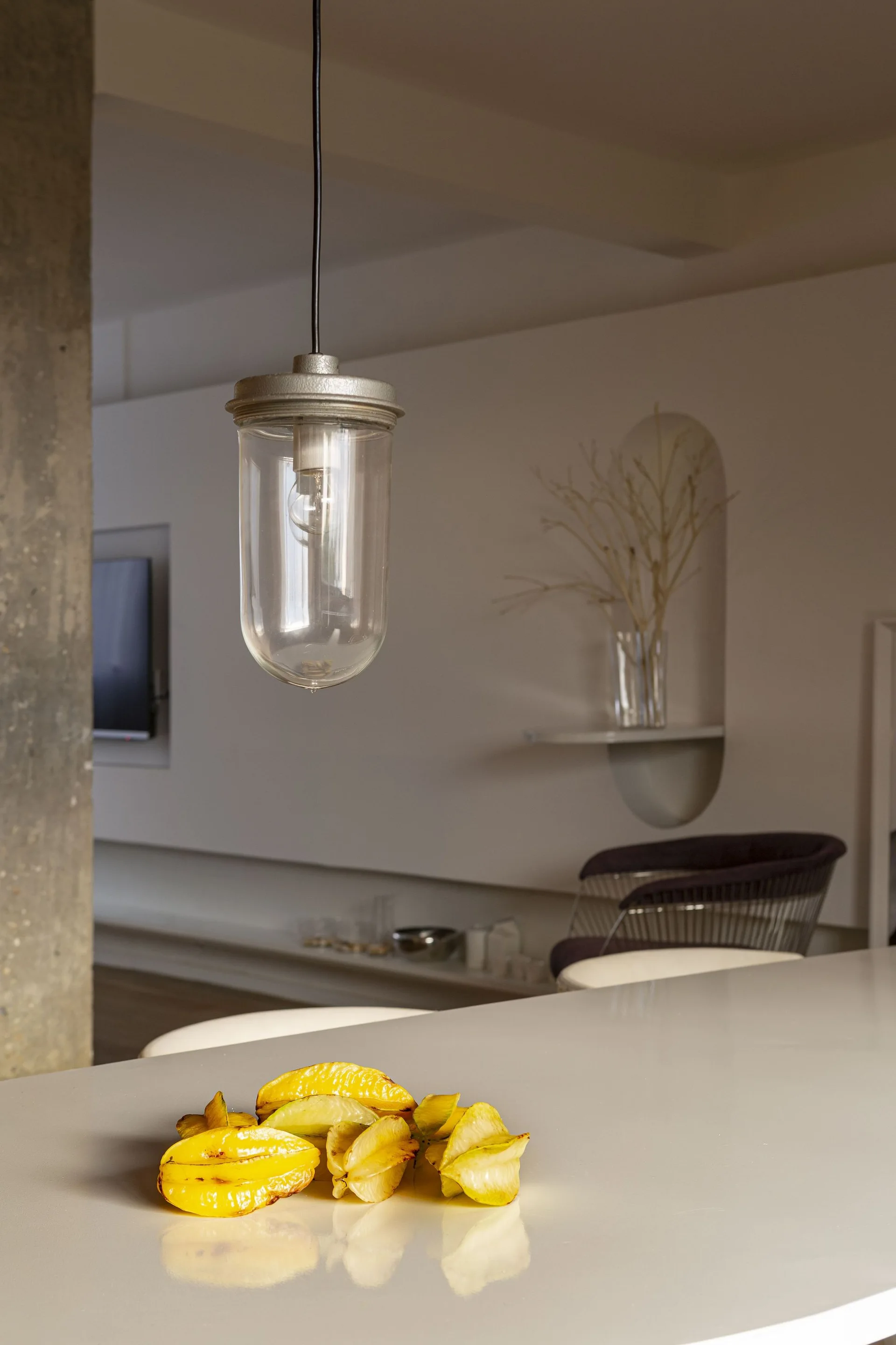 An industrial glass pendant light hangs above a glossy white kitchen counter with star fruit, with a concrete column and arched wall niche in the background.