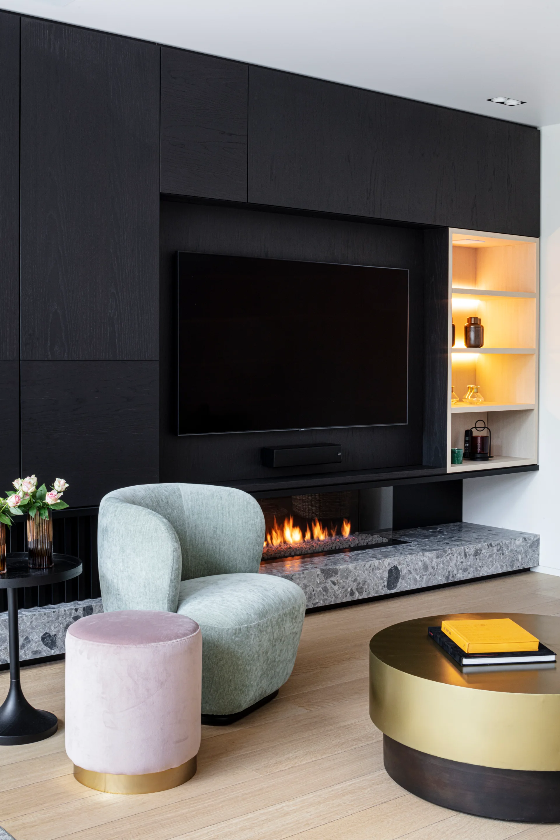 A charcoal wood built-in wall unit features a television, gas fireplace, speckled stone hearth, and a light green armchair with a pink velvet ottoman.