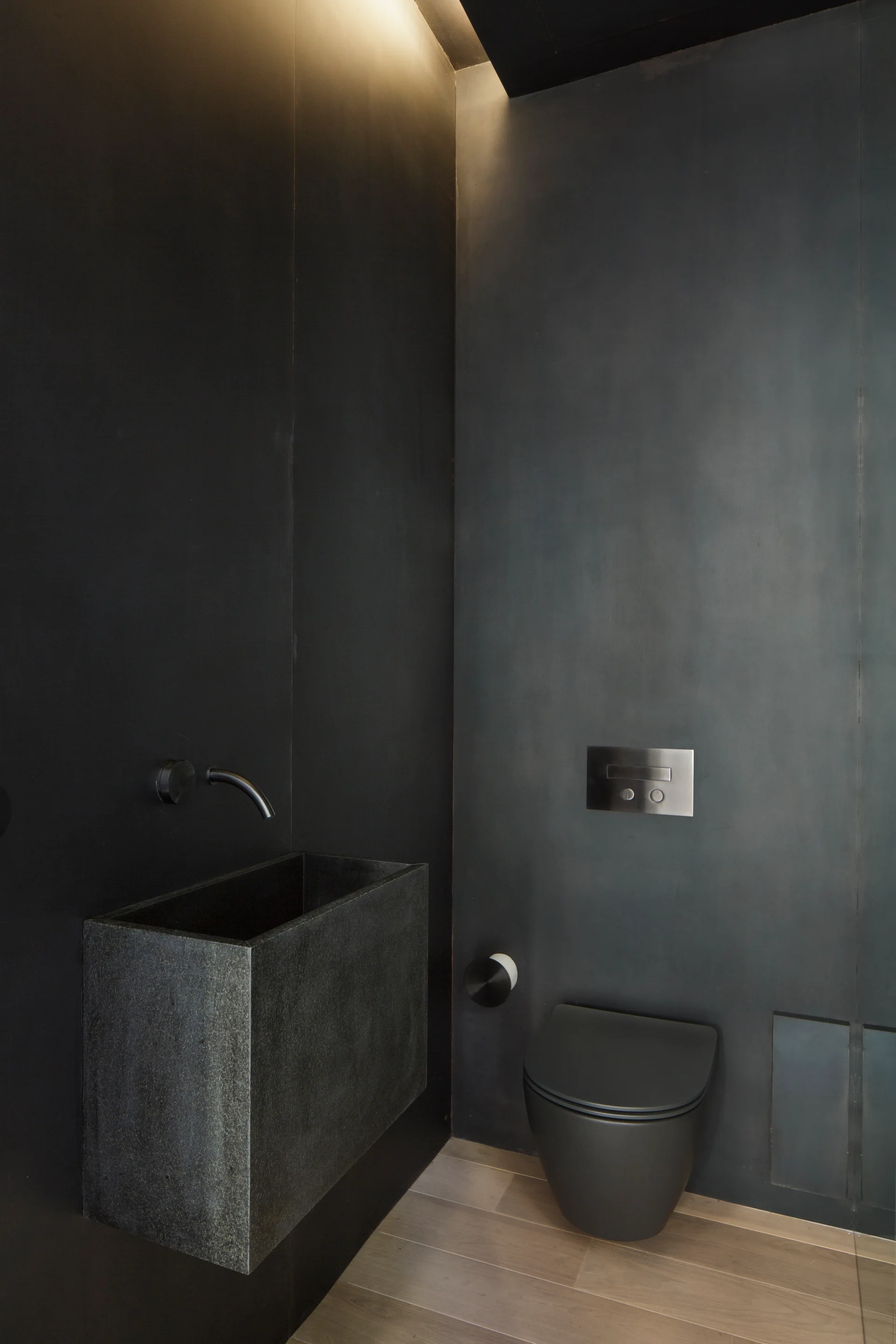 A minimalist dark powder room with matte charcoal walls, a wall-mounted rectangular stone sink, and a black toilet on light wood flooring.