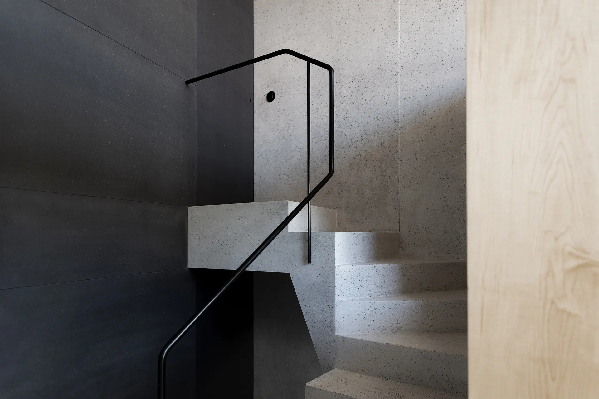 A minimalist stone-like staircase with light speckled treads features a thin black handrail against contrasting matte black and light grey walls.