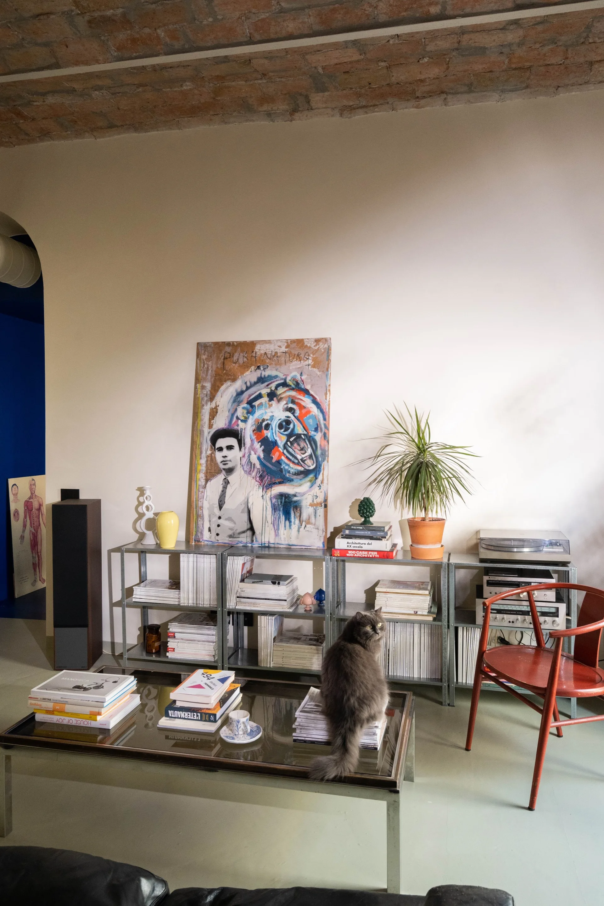 An eclectic living room features a low modular metal shelving unit filled with books under an exposed brick ceiling, next to a red wooden chair.