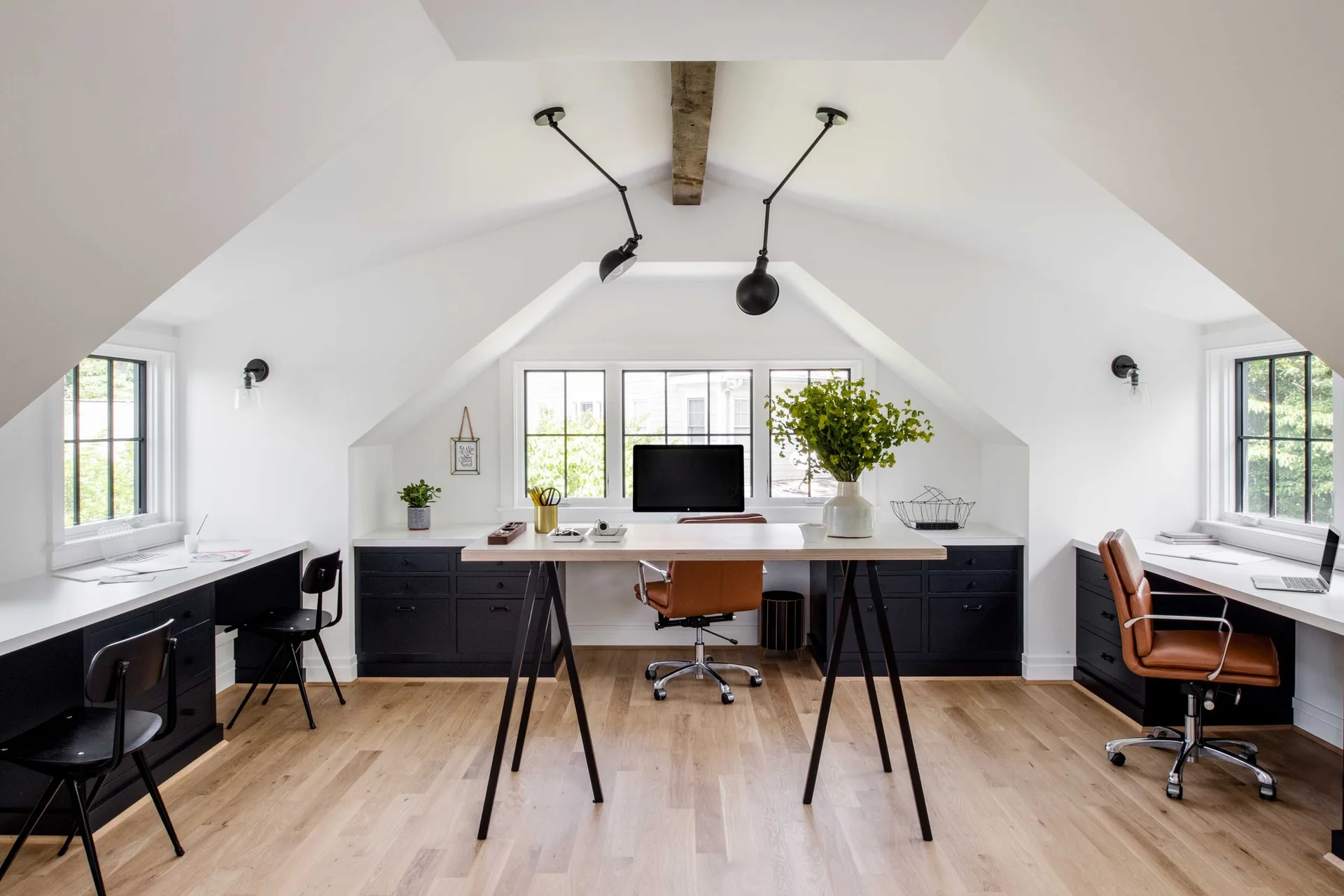 A symmetrical attic home office with a central light wood trestle desk, custom black lower cabinets, and sloped white ceilings.