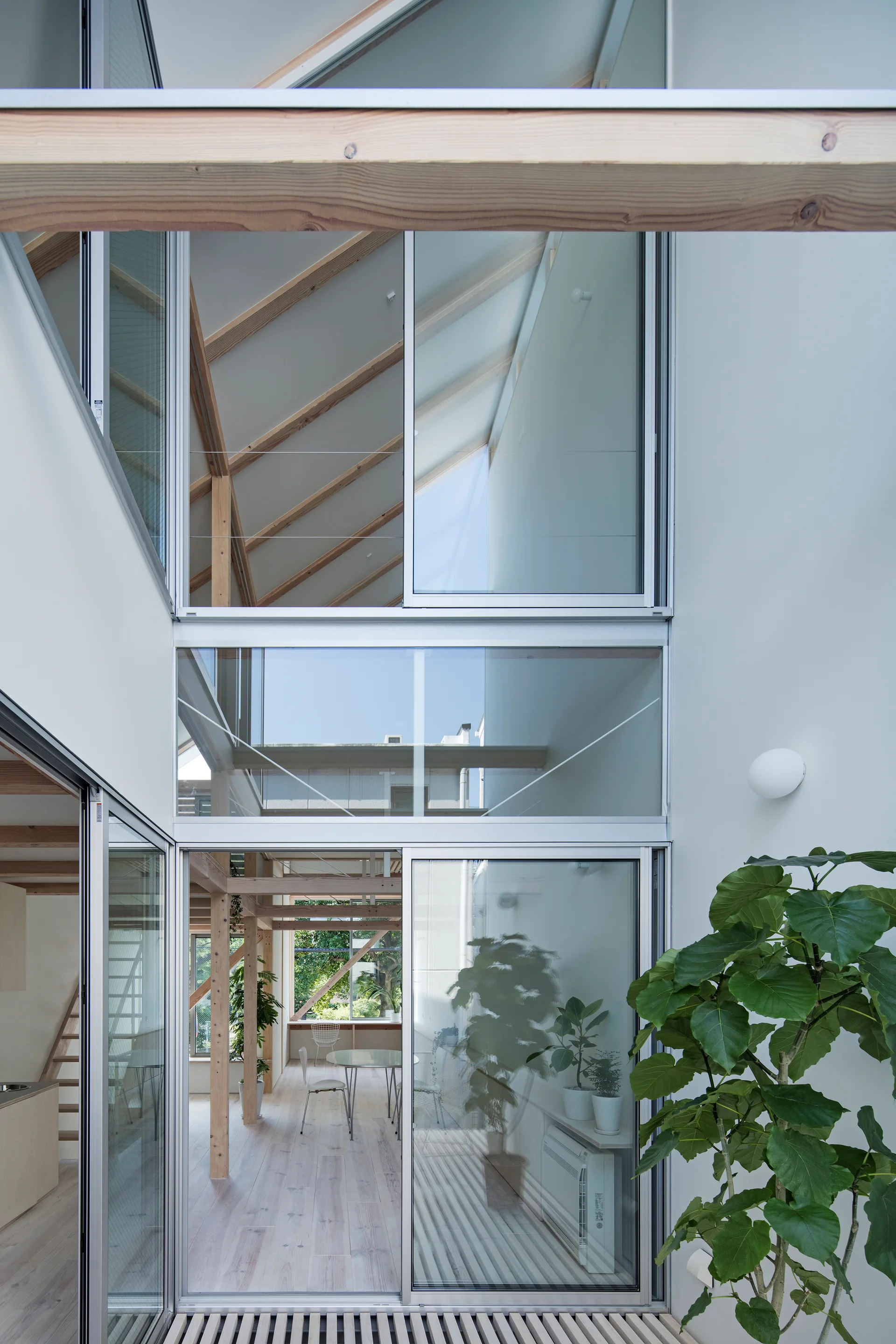 A multi-level modern home interior seen through tall sliding glass doors, featuring exposed wood ceiling beams, white walls, and green potted plants.