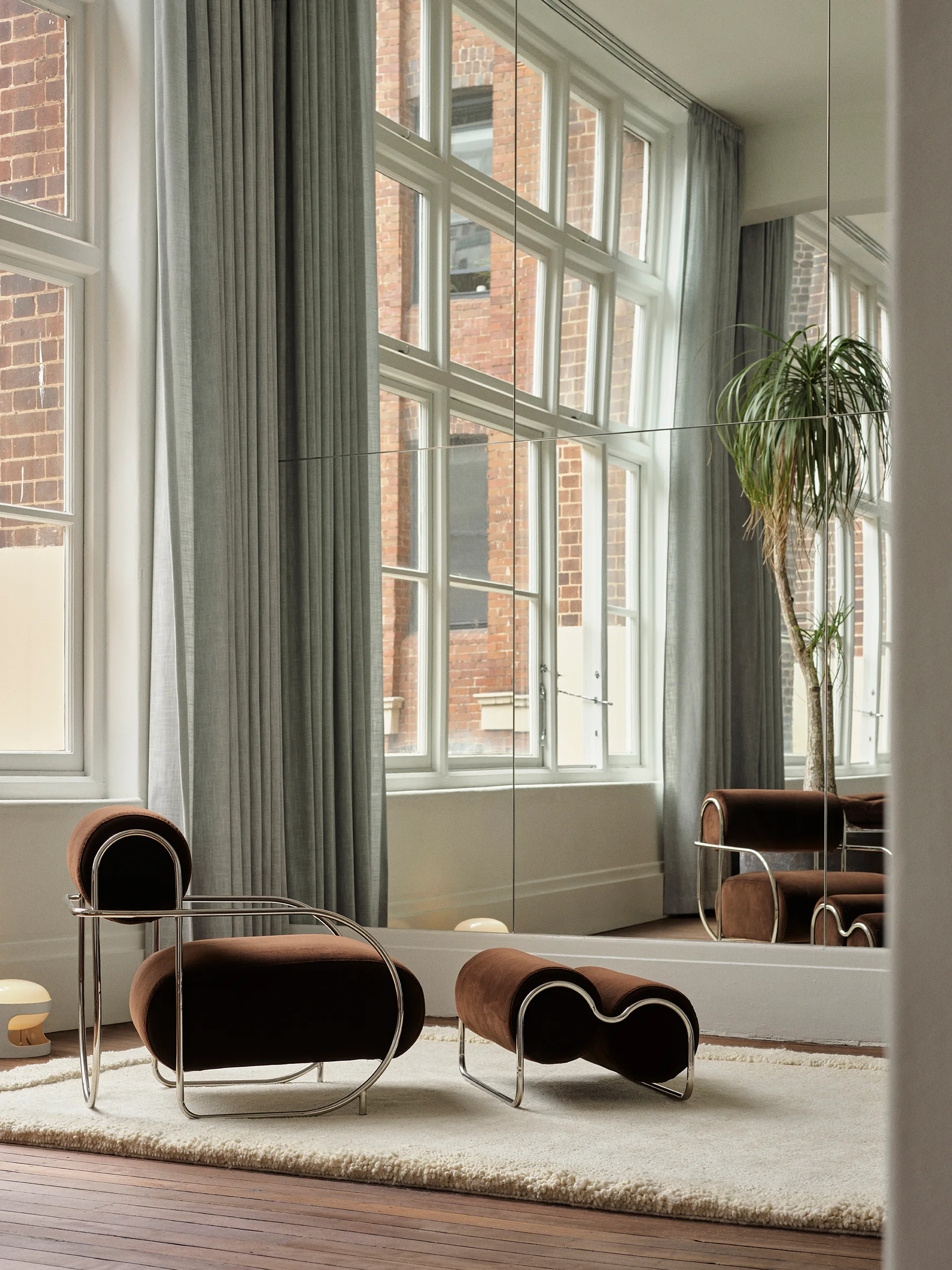 A brown velvet lounge chair and matching ottoman sit on a cream textured rug in front of a large mirrored wall reflecting a window.
