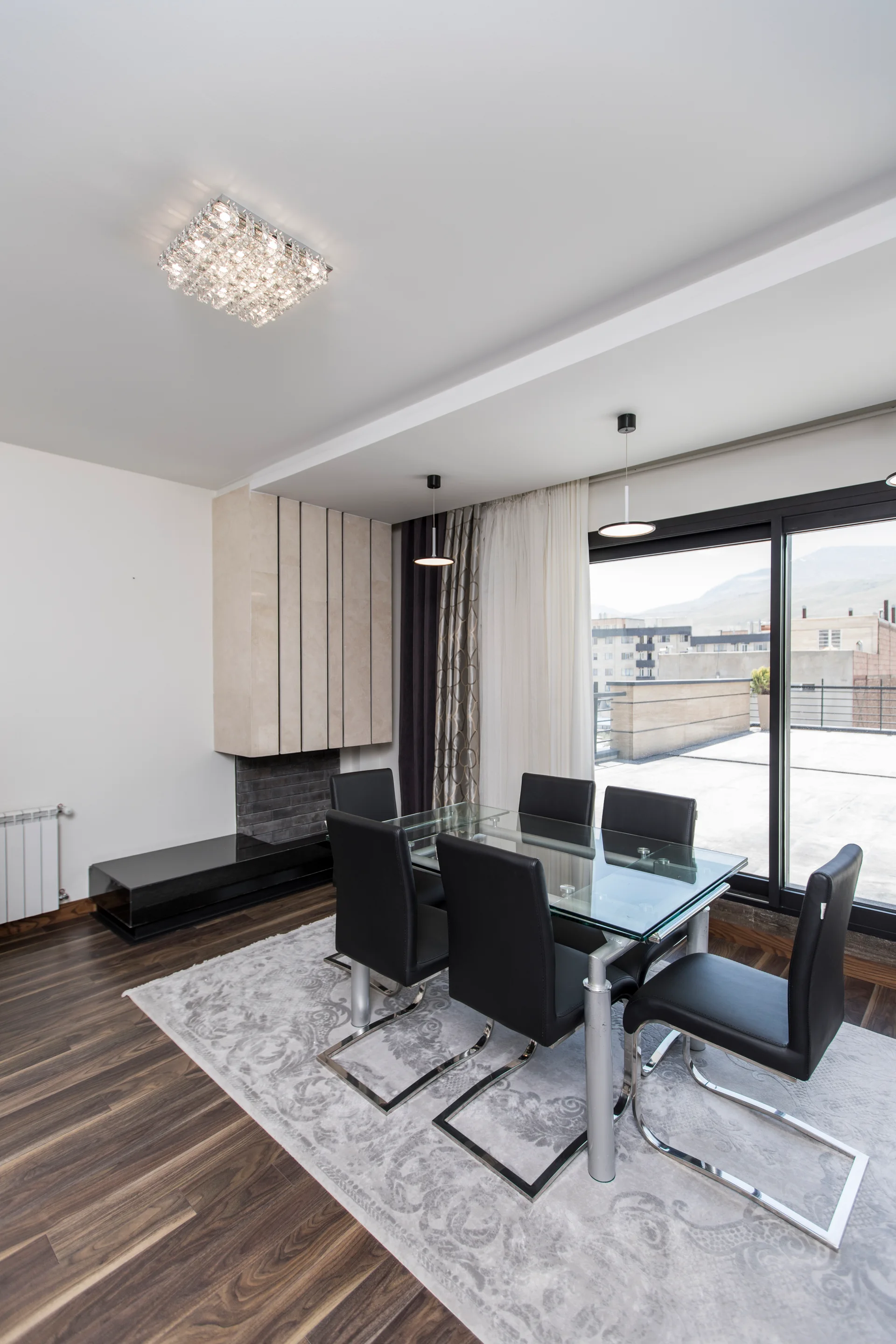 A modern dining room features a glass-top table with black metal-framed chairs on a patterned grey rug next to a stone fireplace.