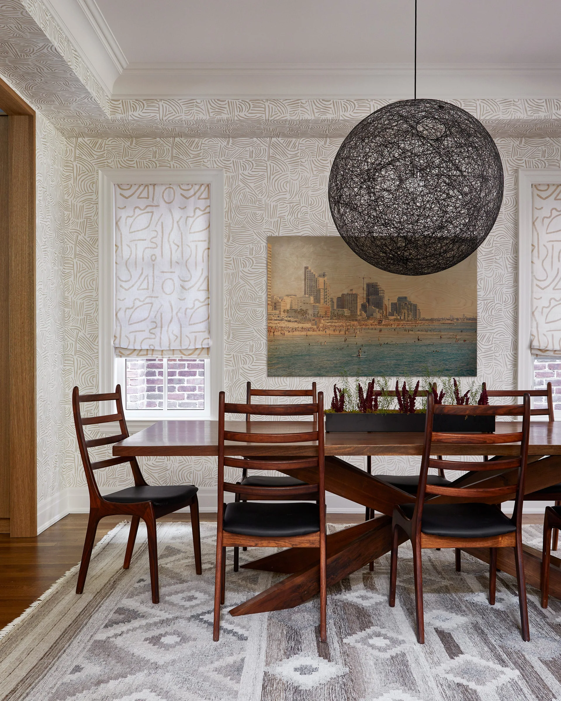 A mid-century modern dining room features a dark wood trestle table, matching chairs, a large wire sphere pendant light, and beige patterned wallpaper.