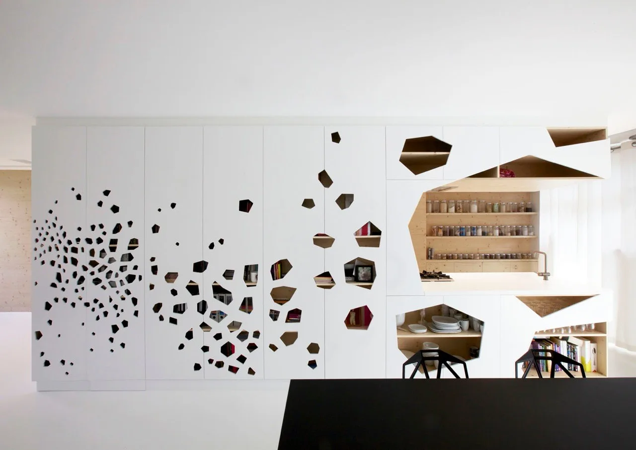 Floor-to-ceiling white kitchen cabinetry featuring unique geometric cutouts that reveal wood-toned open shelves filled with dishes, books, and jars.