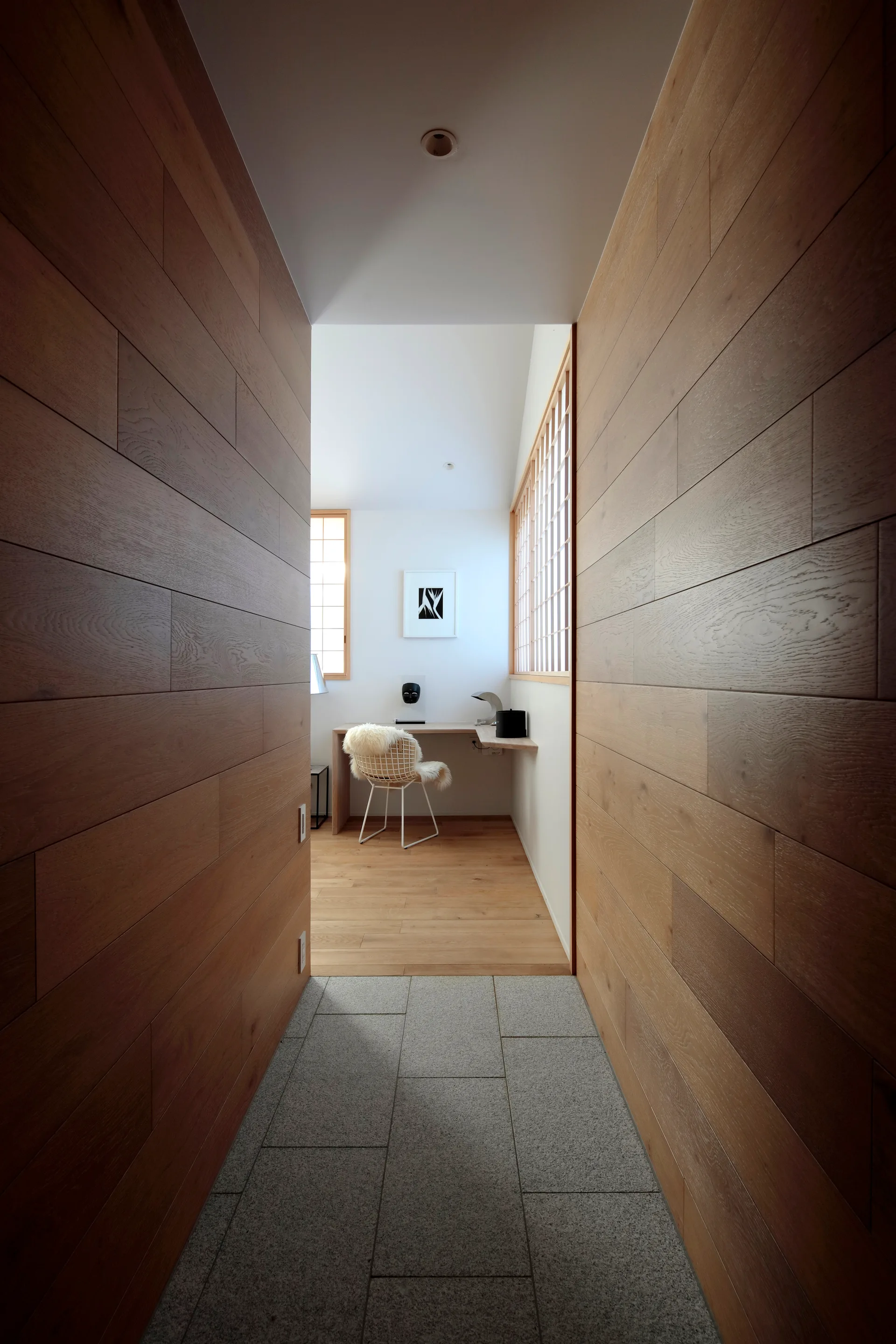 A perspective view down a wood-paneled hallway with stone flooring toward a minimalist home office featuring a floating desk and shoji window screens.