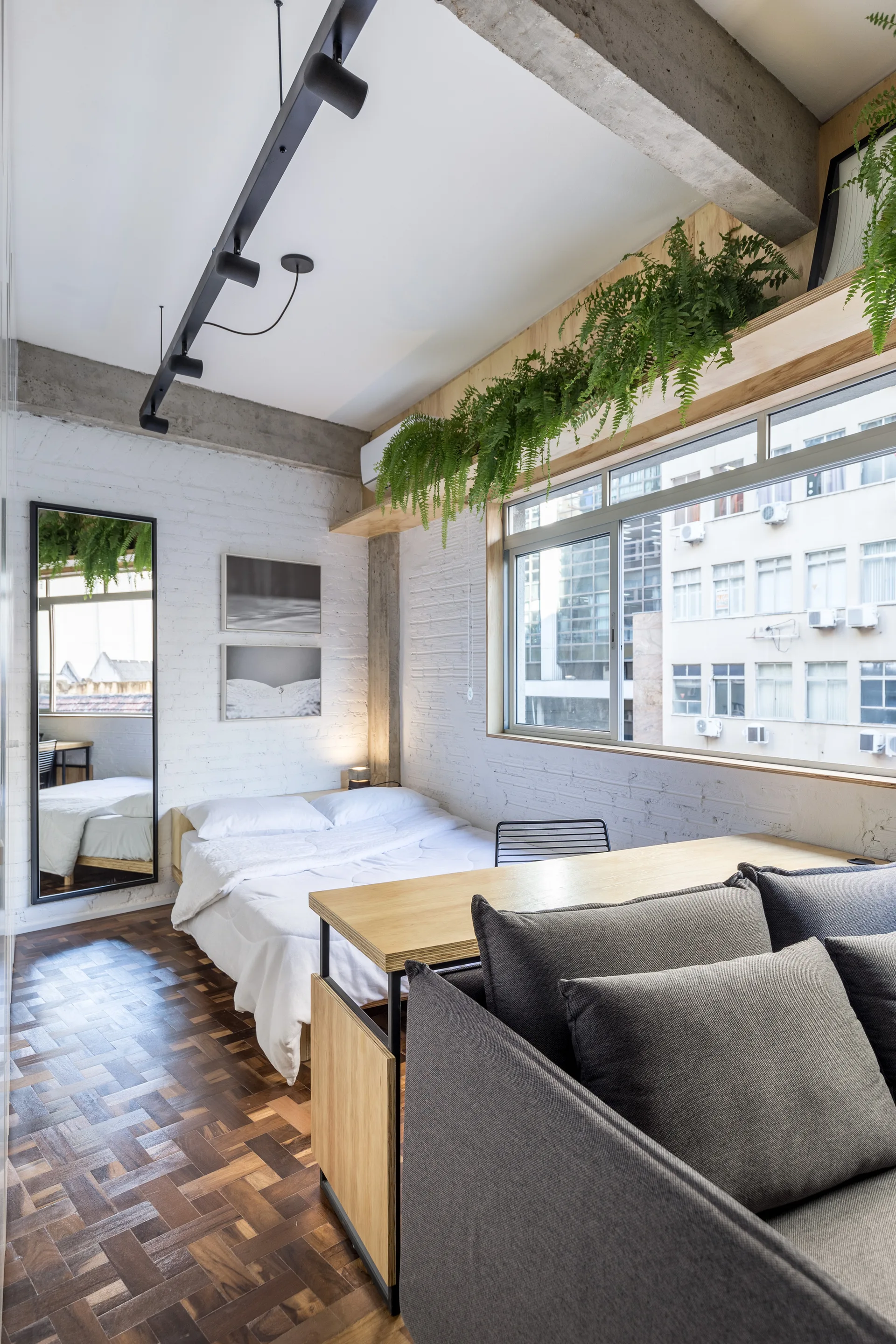 A modern bedroom featuring a white bed, painted brick walls, dark parquet wood floors, and a light wood shelf with green plants above a window.