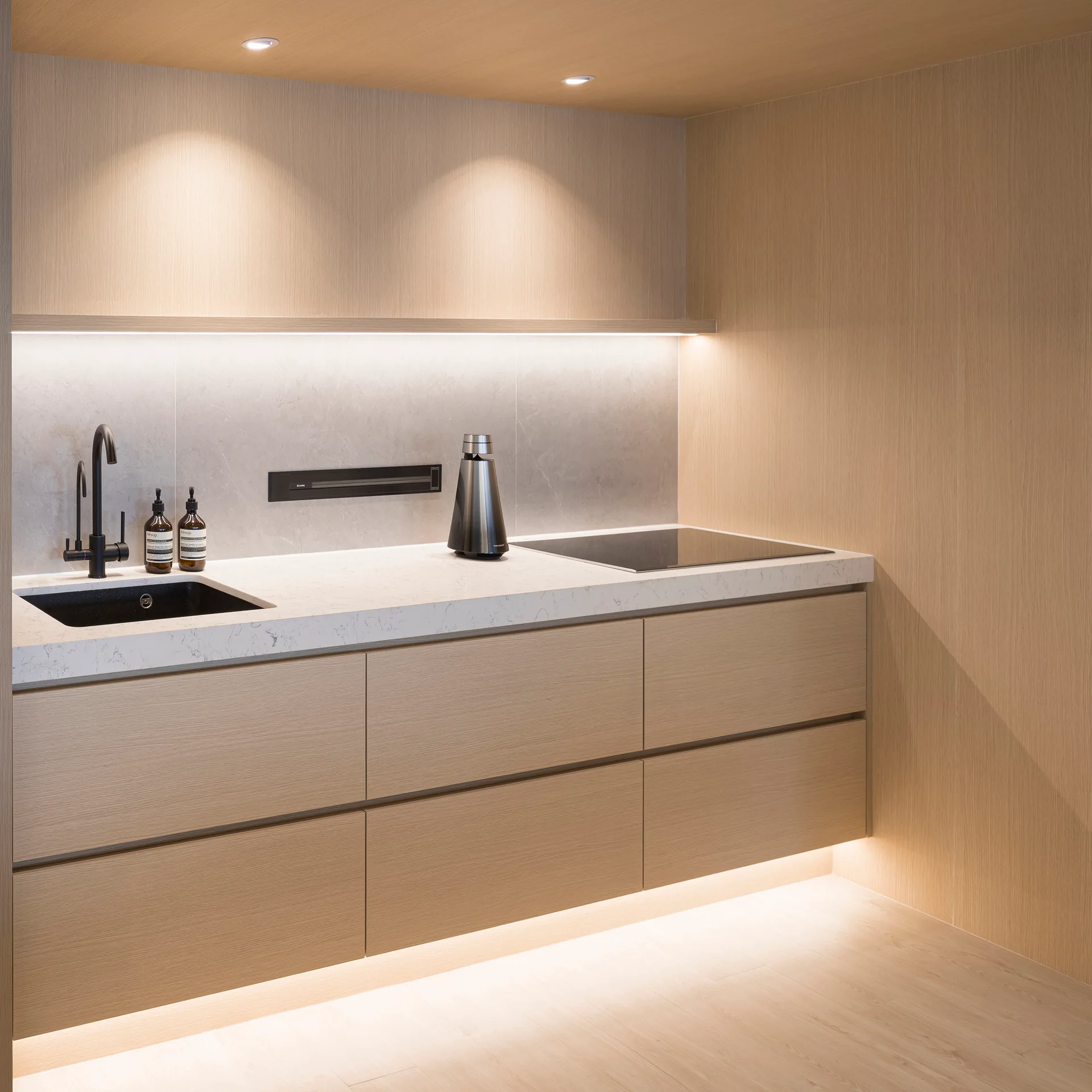 A minimalist wood-paneled kitchen features flat-front cabinets, a white stone countertop with a black undermount sink, and warm integrated LED strip lighting.