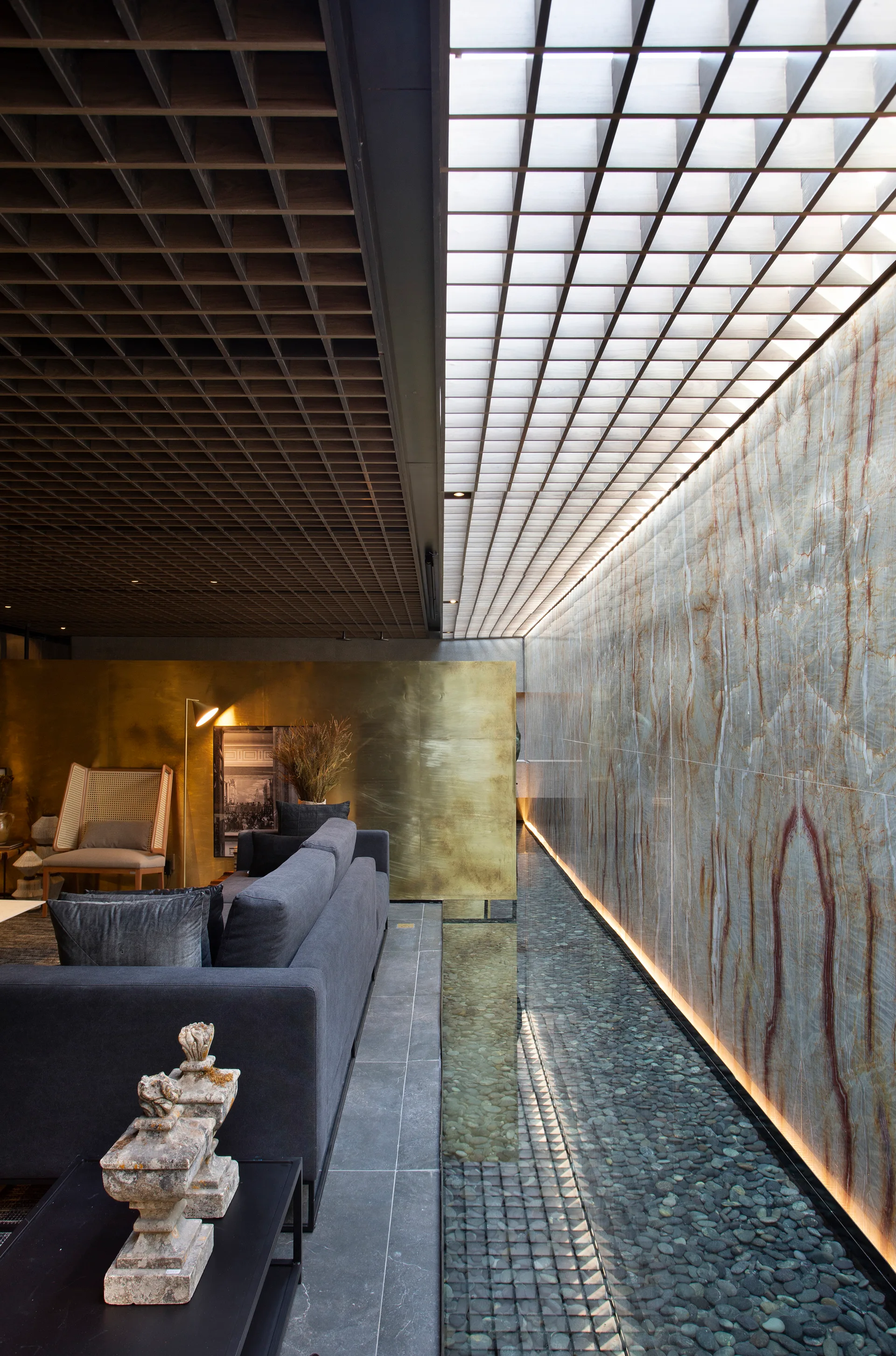 A dark grey sofa sits next to an indoor pebble-filled water feature beneath a textured stone wall and a wood grid ceiling.