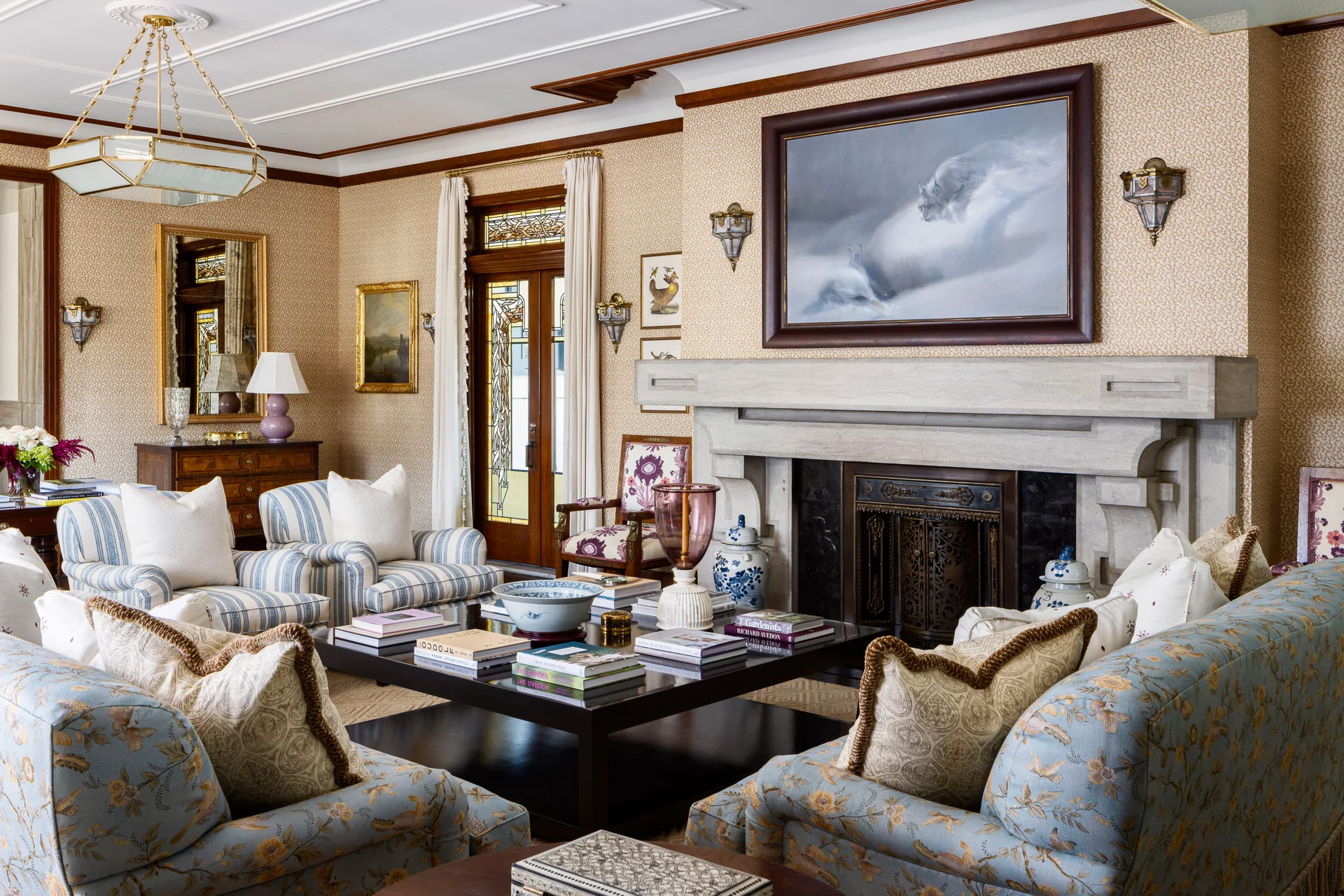 A formal traditional living room features blue patterned sofas, a large stone fireplace with a framed painting above, and stained glass doors.