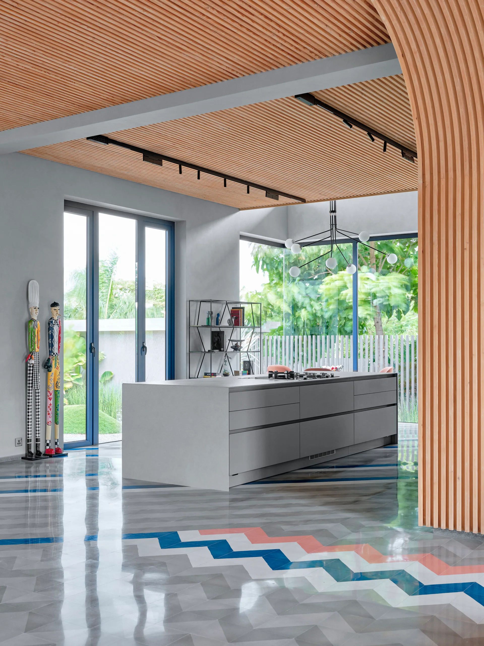A modern kitchen features a grey island under a slatted wood ceiling, with a glossy chevron patterned floor and large floor-to-ceiling windows.