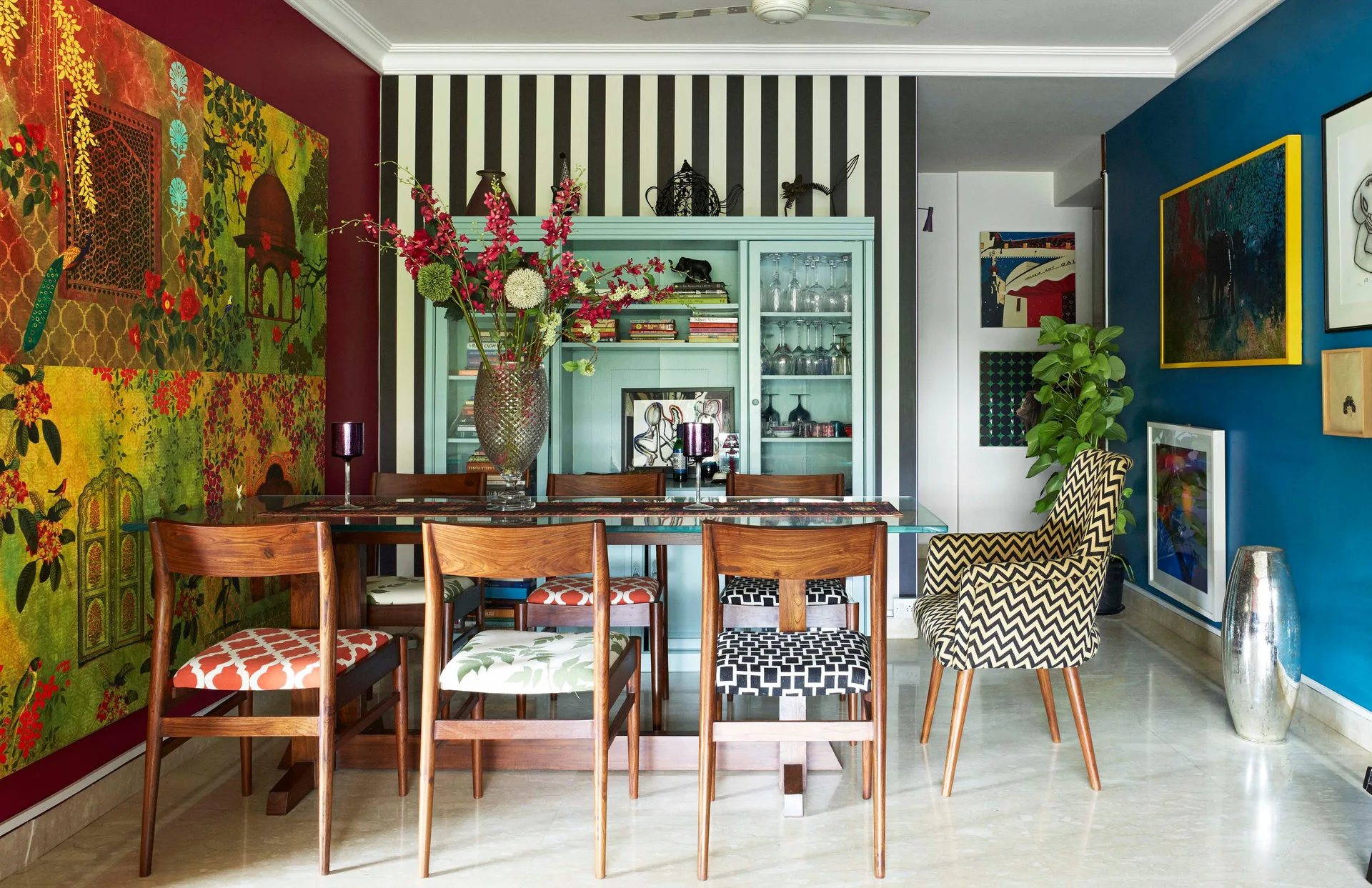 An eclectic dining room featuring a glass-topped wooden table, mismatched patterned chairs, and a mint green cabinet against striped wallpaper.