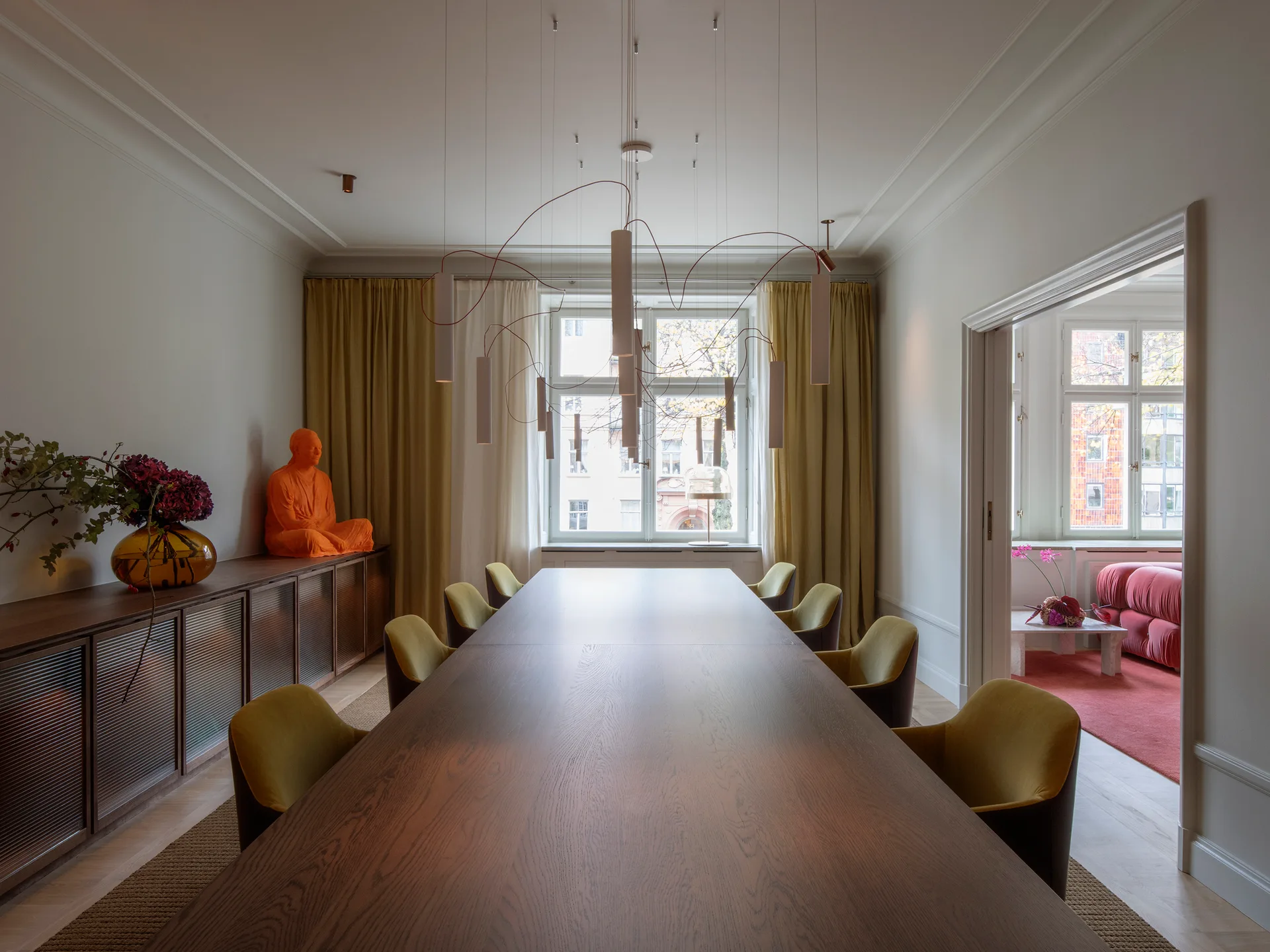 A long dark wood dining table surrounded by mustard yellow chairs beneath a large sculptural white pendant light in a neutral-toned room.