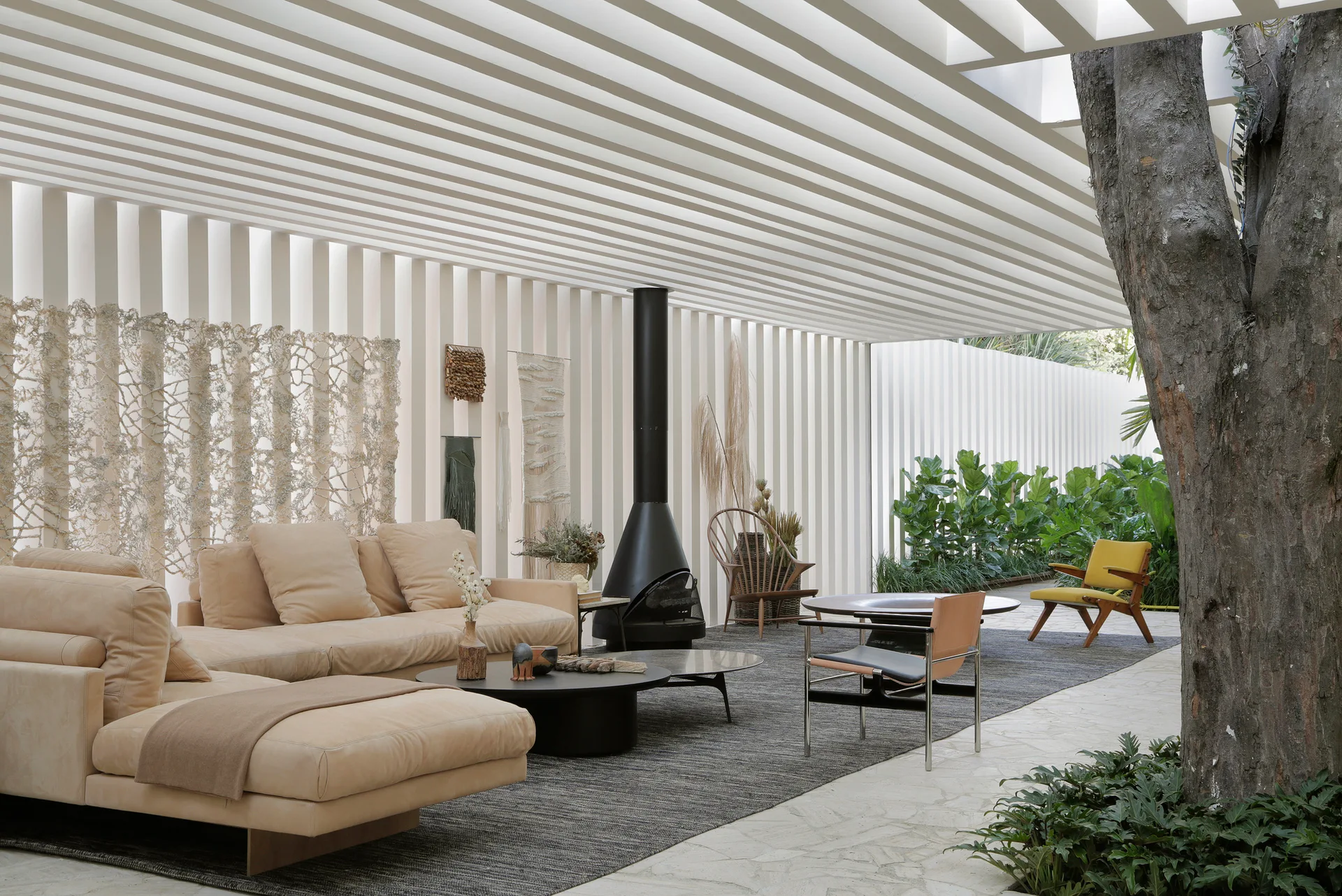 A covered outdoor patio featuring a beige sectional sofa, a freestanding black metal fireplace, and a large tree growing through the stone tile floor.