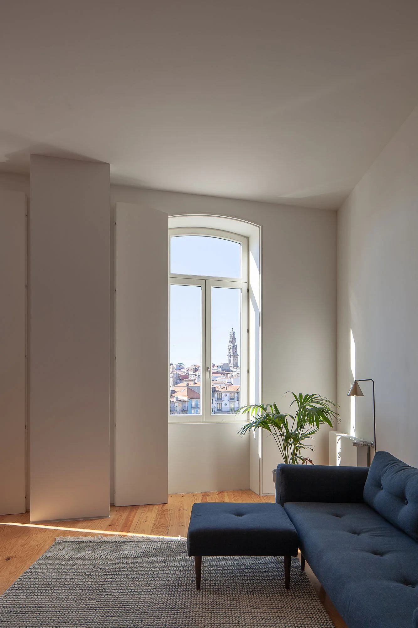 A dark blue sofa sits on a textured grey rug next to a tall arched window offering a view of a historic cityscape.