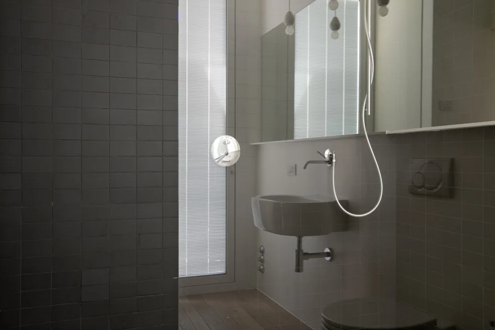 A modern minimalist bathroom featuring a wall-mounted porcelain sink, chrome plumbing, dark grey square wall tiles, and wood floors.