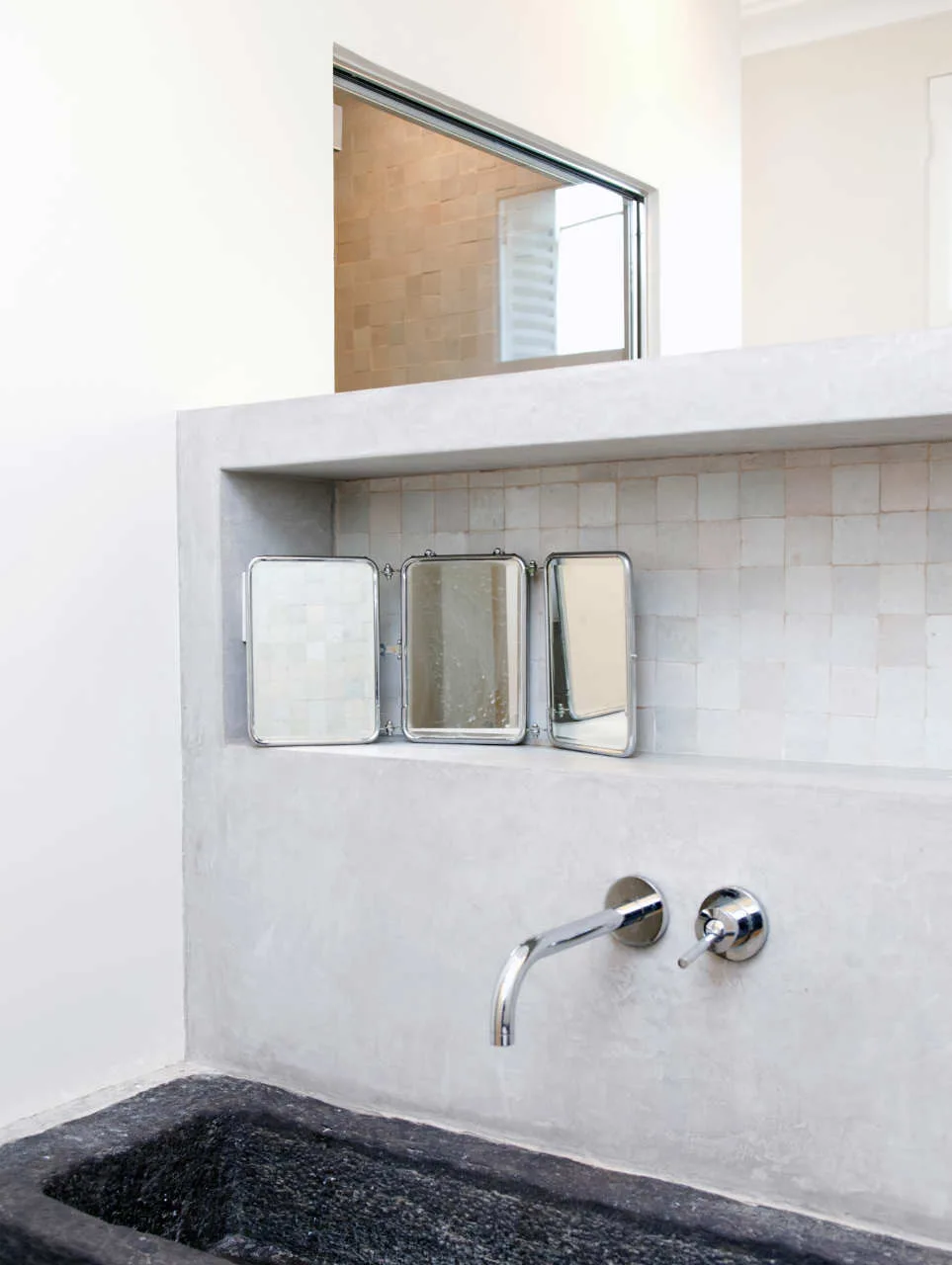 A grey concrete bathroom vanity features a wall-mounted chrome faucet below a tiled niche containing a small three-panel folding triptych mirror.