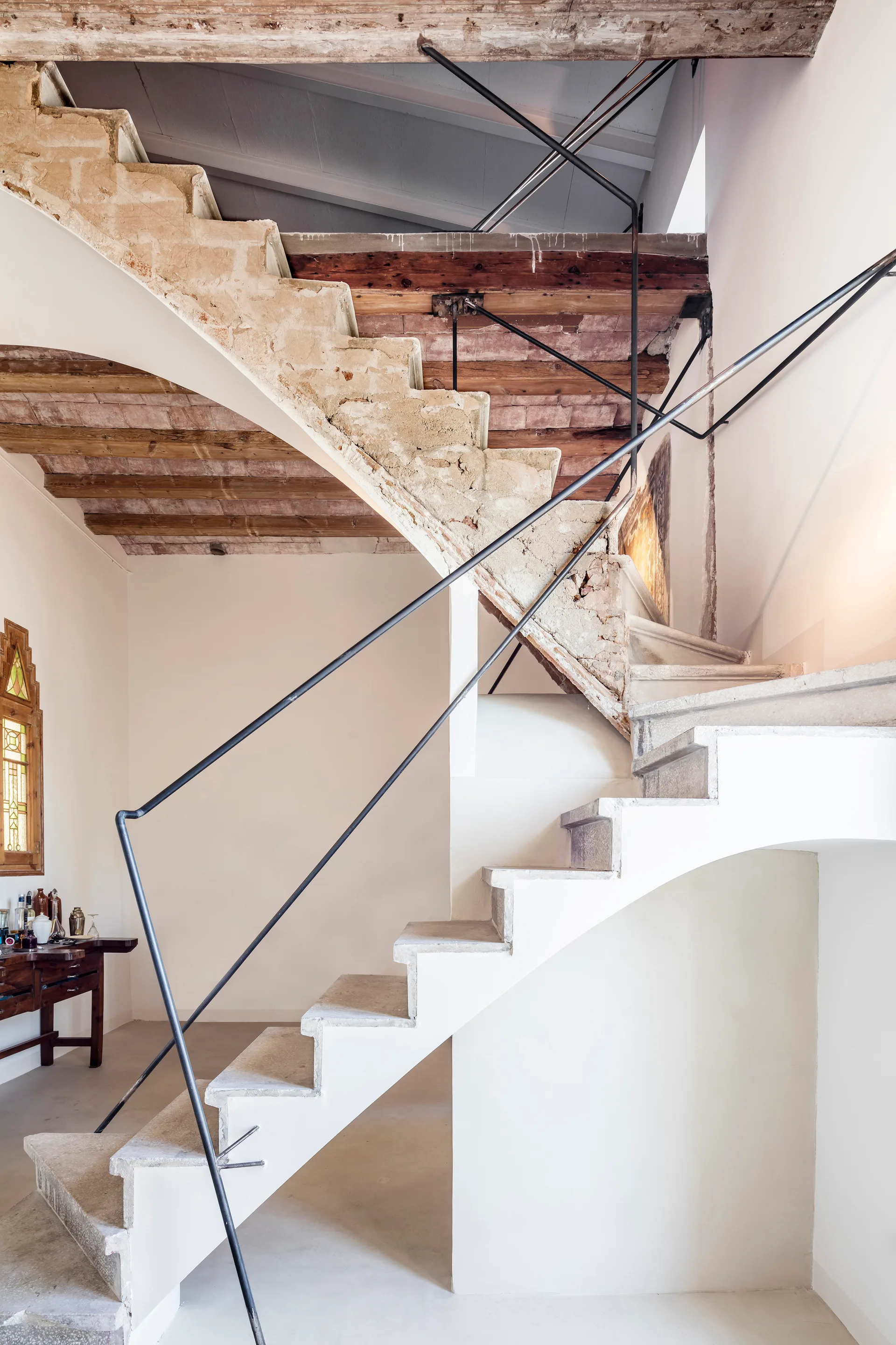 A sculptural concrete staircase with black metal railings features exposed stone underneath and rustic wood ceiling beams.
