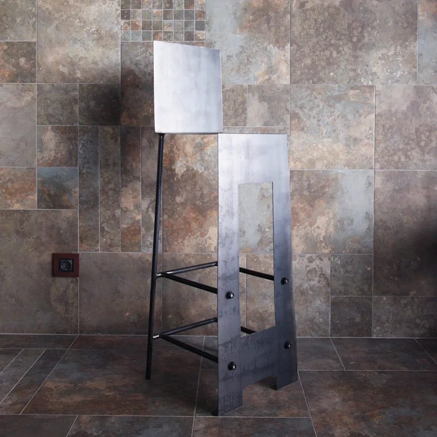 A minimalist industrial metal bar stool with an angular seat stands against a brown and grey slate tile wall.