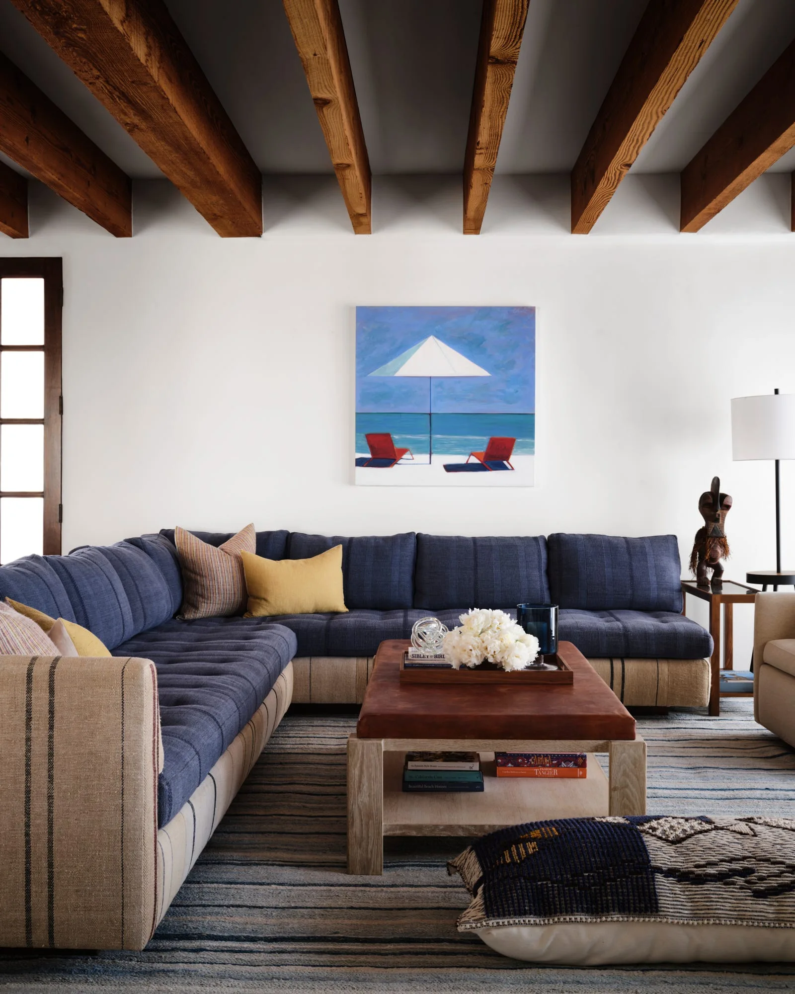A blue sectional sofa sits under exposed wood ceiling beams in a living room with a leather-topped coffee table and a striped rug.
