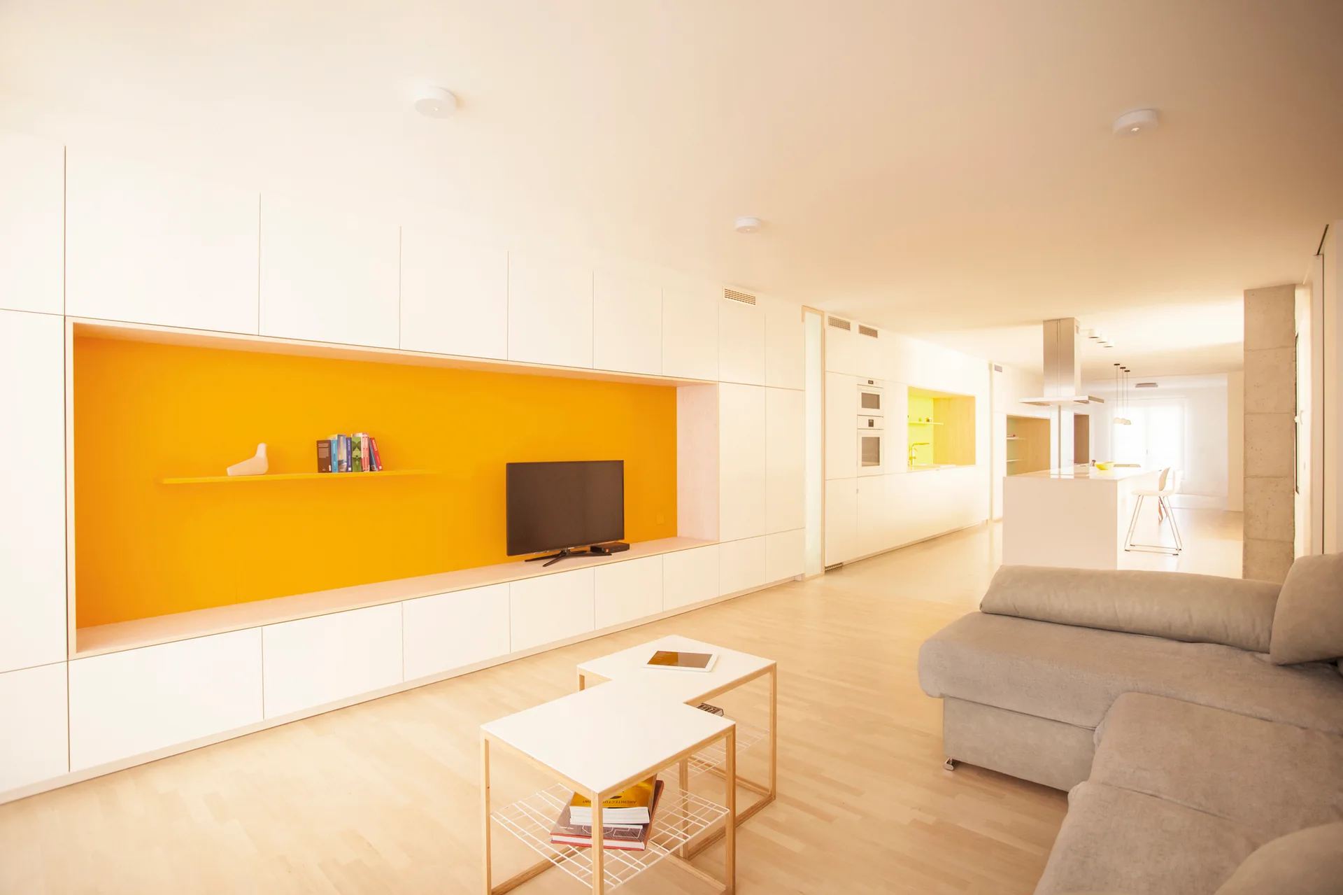 A modern open-plan living room with a large white built-in wall unit featuring a bright yellow recessed shelf, a beige sofa, and light wood flooring.