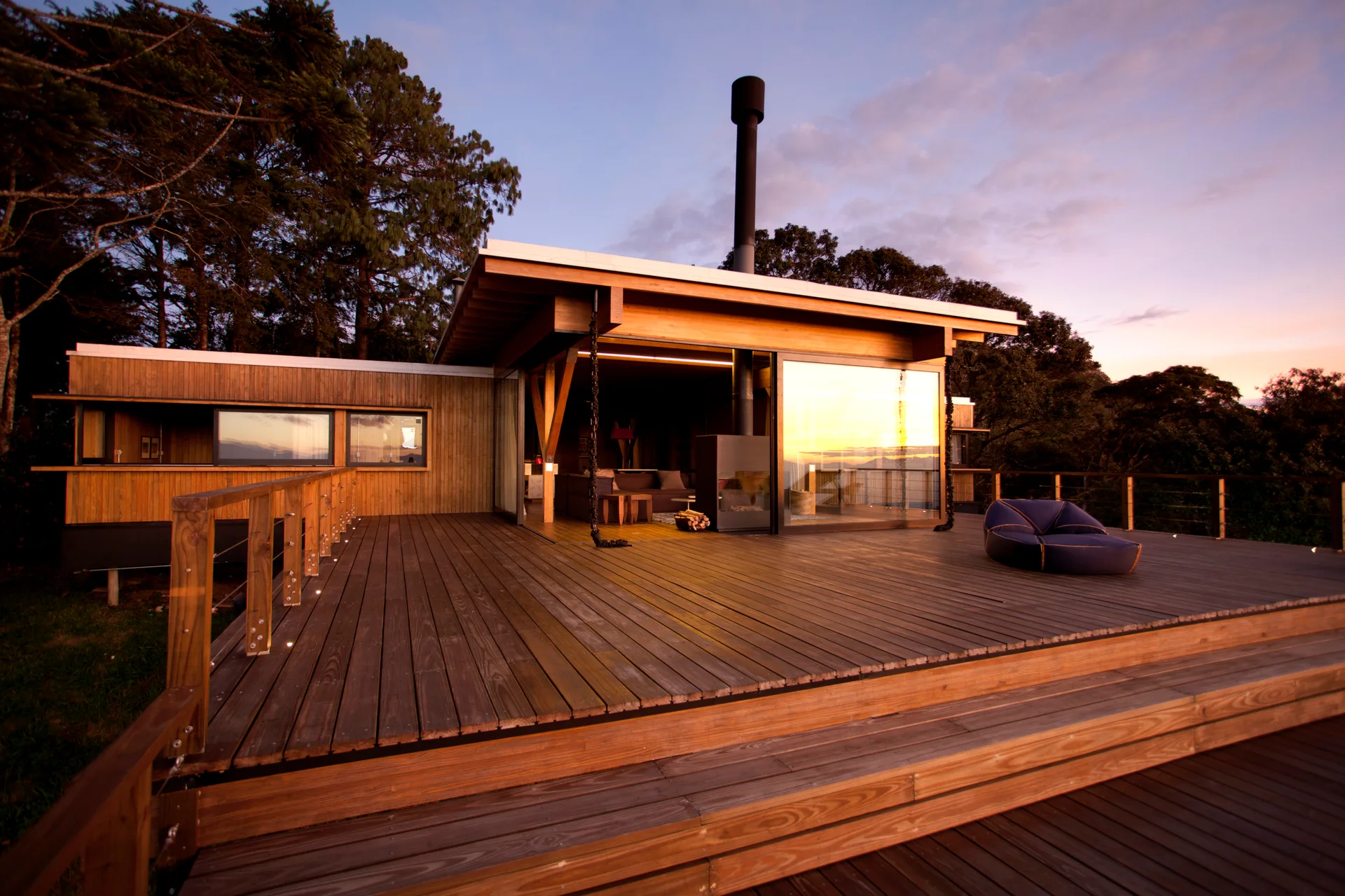 A modern wood clad cabin features a large multi level wooden deck with steps, expansive glass sliding doors, and a black chimney pipe at twilight.