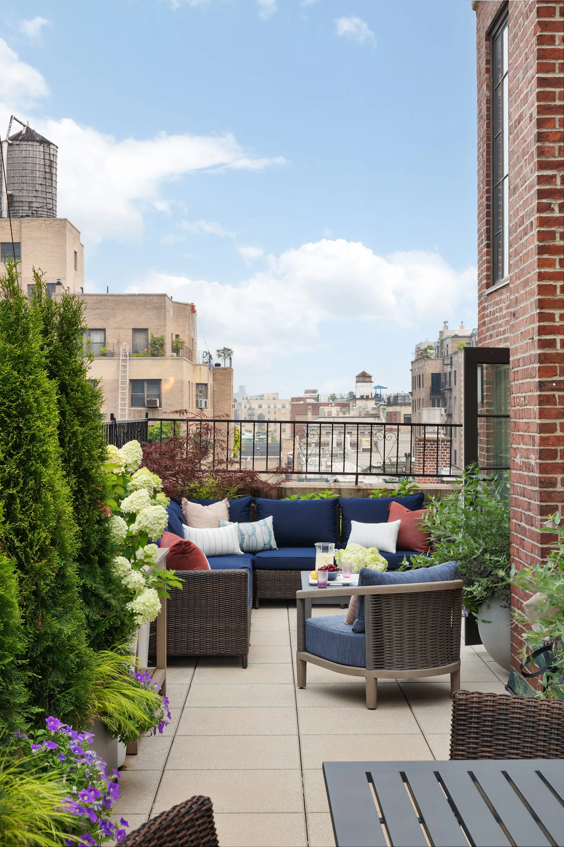 A rooftop terrace features a woven wicker sectional sofa with navy cushions and accent pillows next to potted evergreens and a brick wall.