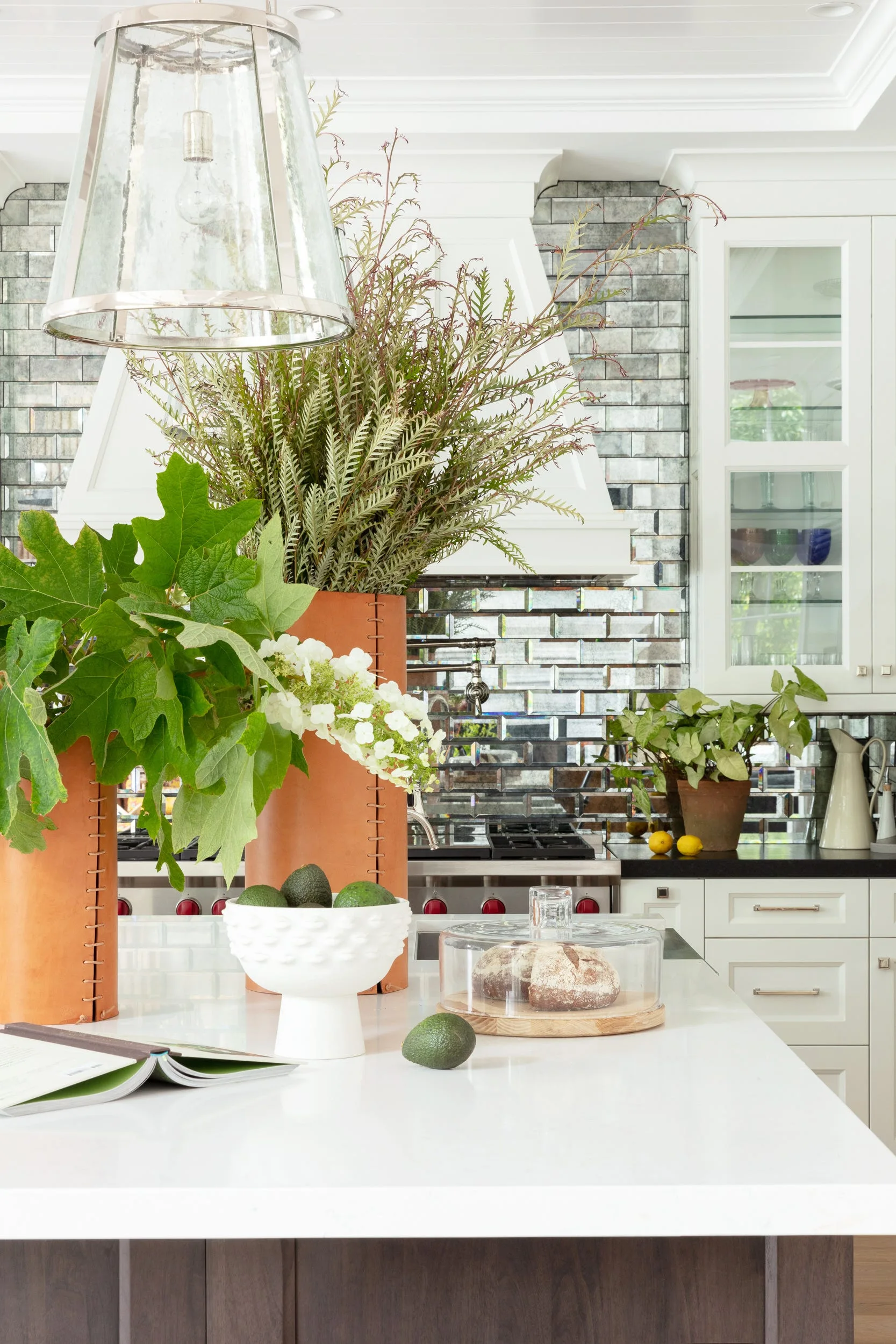 A white kitchen island features tall leather-wrapped vases with greenery in front of a mirrored subway tile backsplash and white cabinets.