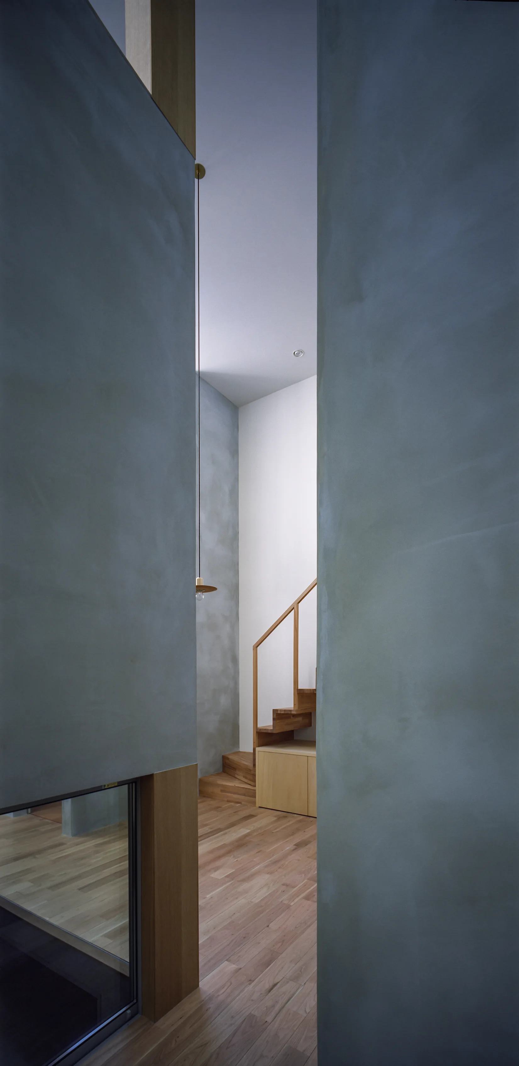A modern wooden staircase with a simple handrail is seen through a narrow vertical opening framed by tall matte grey plaster walls.