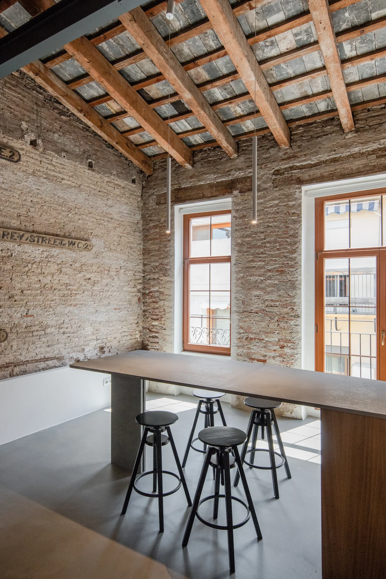 An industrial dining area features a long gray table and black swivel stools set against exposed brick walls and rustic wooden ceiling beams.