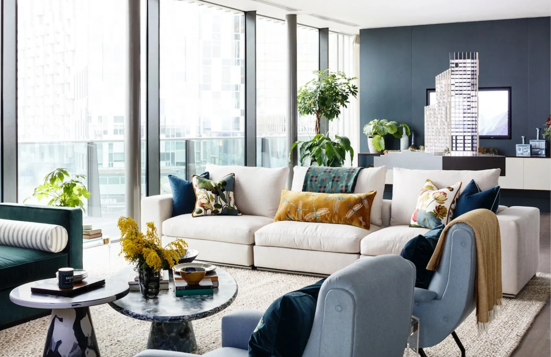 A modern living room featuring a cream sectional sofa, light blue armchairs, and a round stone coffee table set against floor-to-ceiling windows.
