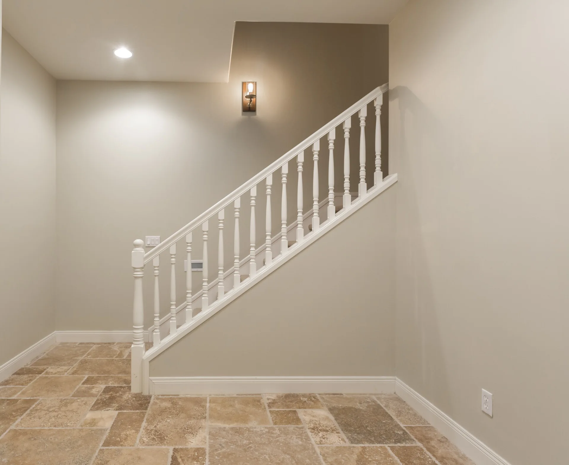 A staircase with a white wooden railing and balusters is set against beige walls and textured tan stone tile flooring.
