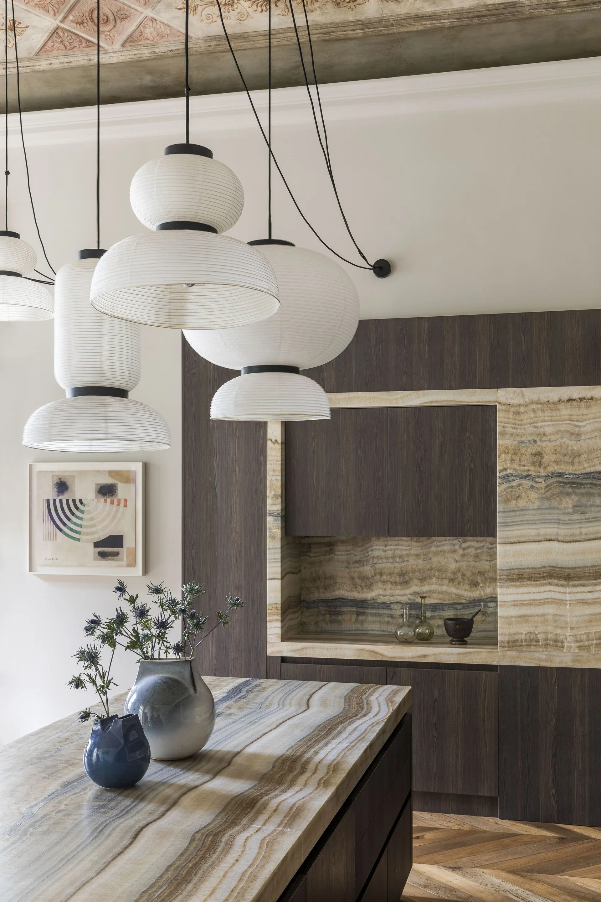 A modern kitchen with a striated stone island, dark wood cabinetry featuring a matching stone niche, and tiered white paper pendant lights.