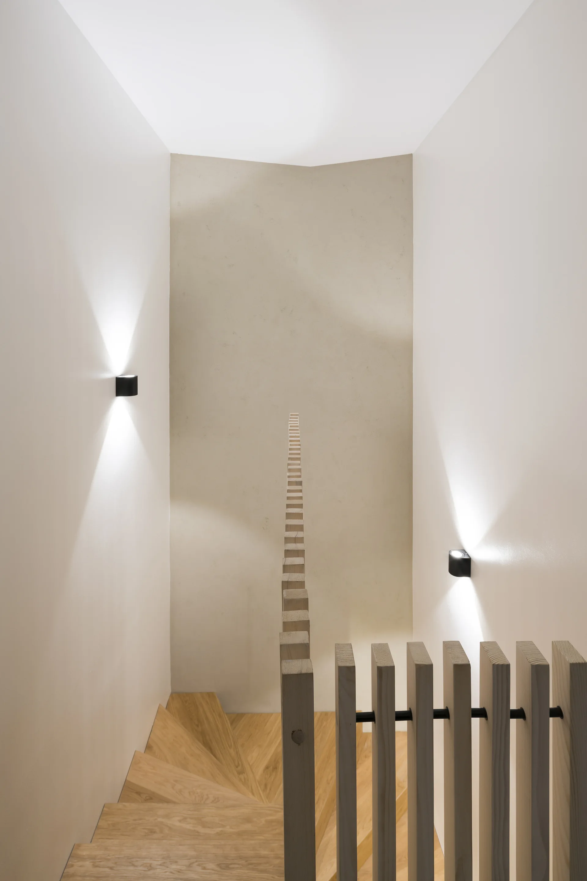 A minimalist wood staircase with winding steps, a vertical slatted railing, and two black wall sconces mounted on off-white walls.